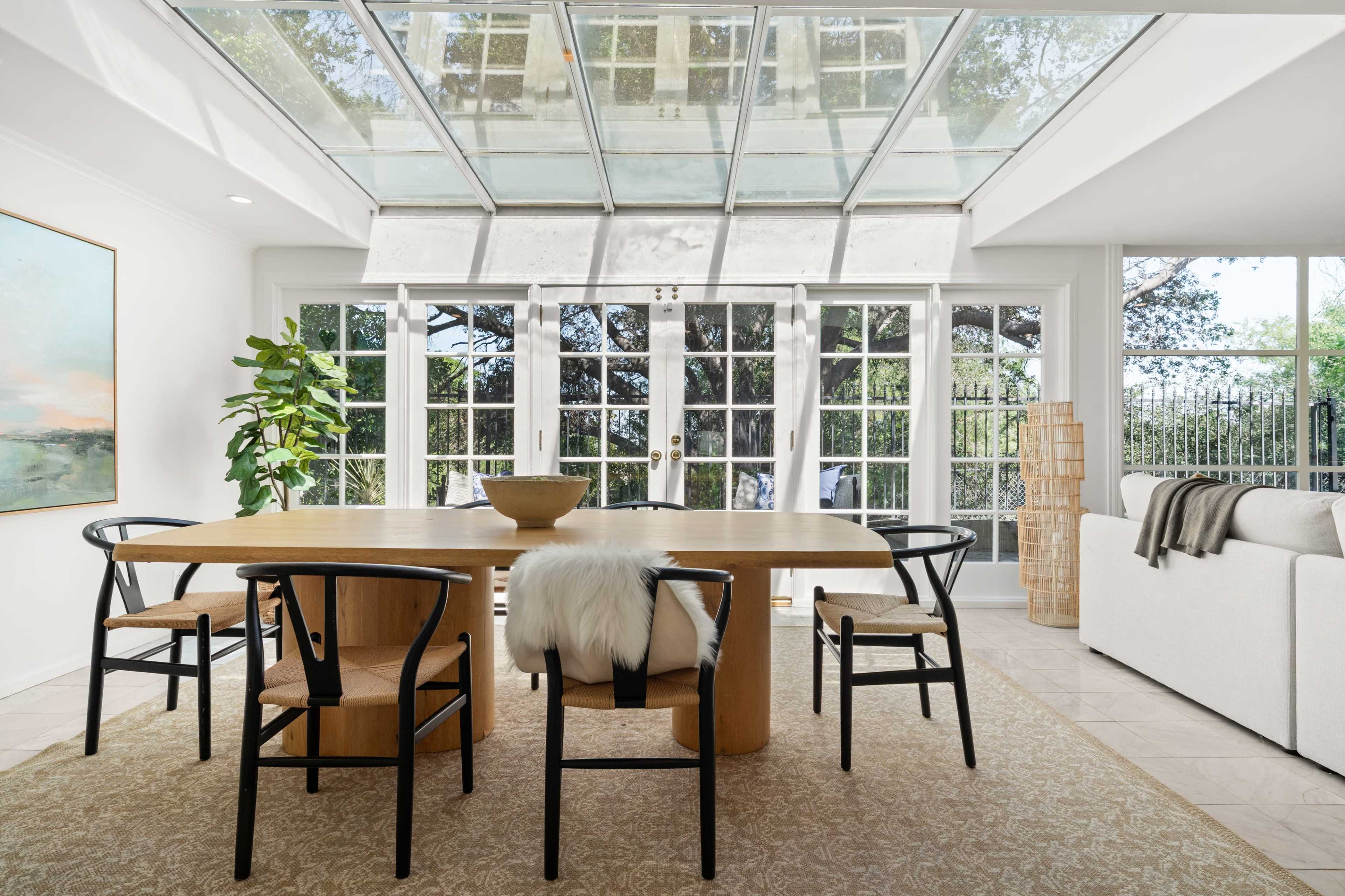 The image shows a bright dining area with a large wooden table surrounded by chairs, a potted plant, and glass doors leading to an outdoor space.