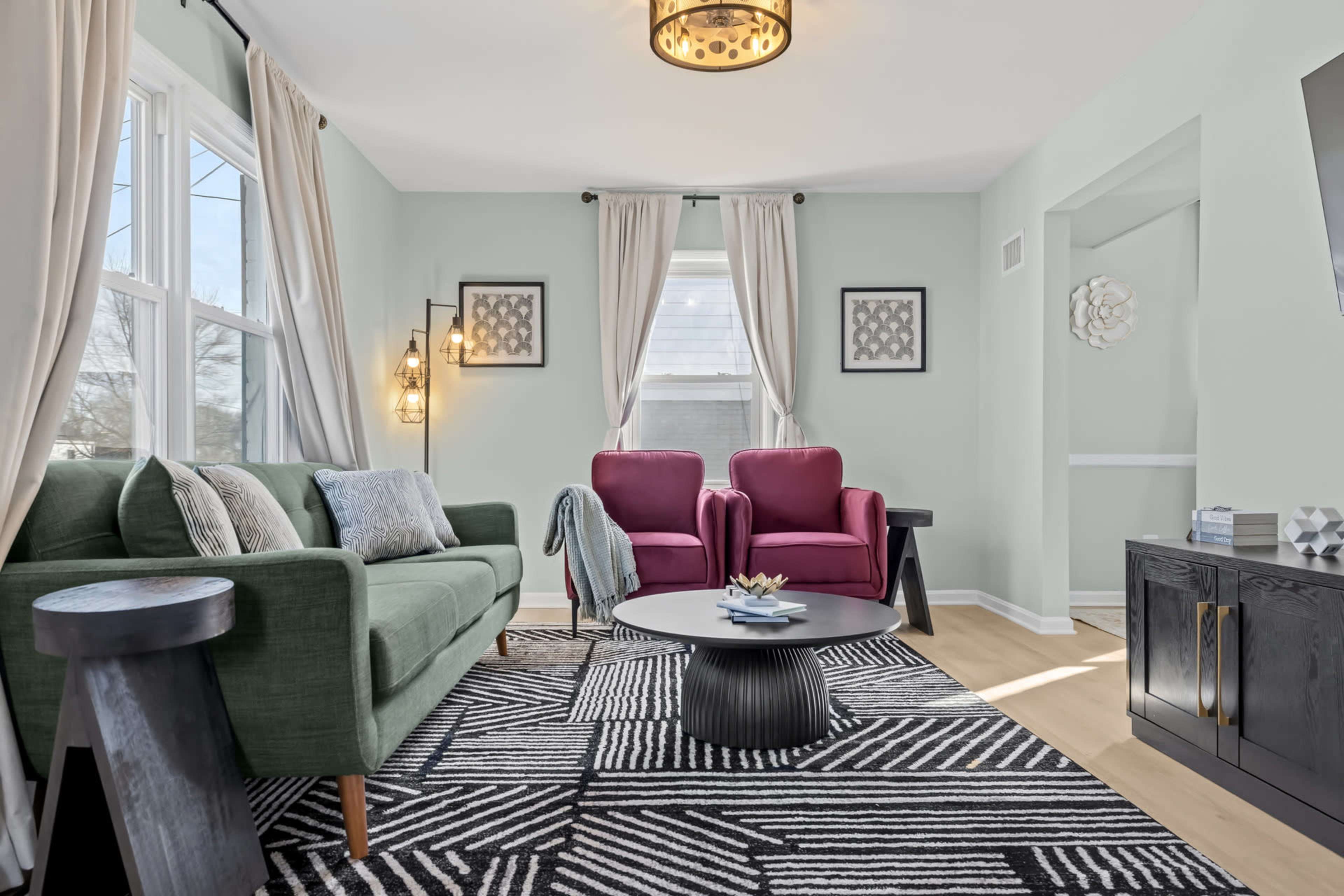 The image shows a light-filled living room featuring a green sofa, two pink chairs, and a round black coffee table on a patterned rug.