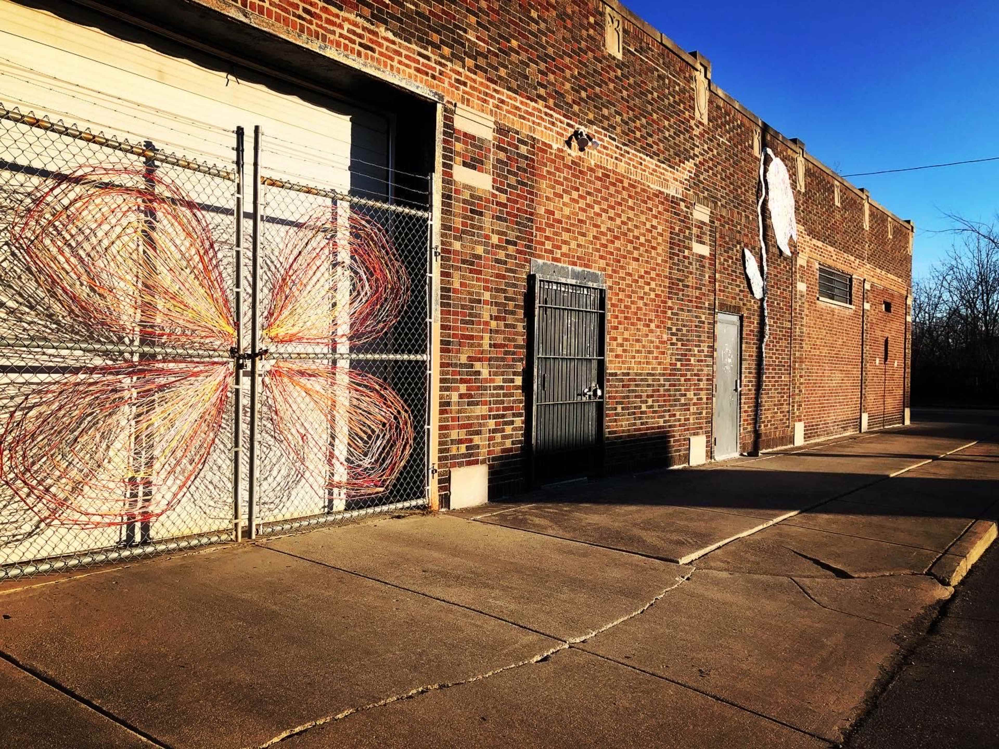 A brick building features a chain-link fence with a colorful mural resembling a flower on one side and a series of doors on the other.