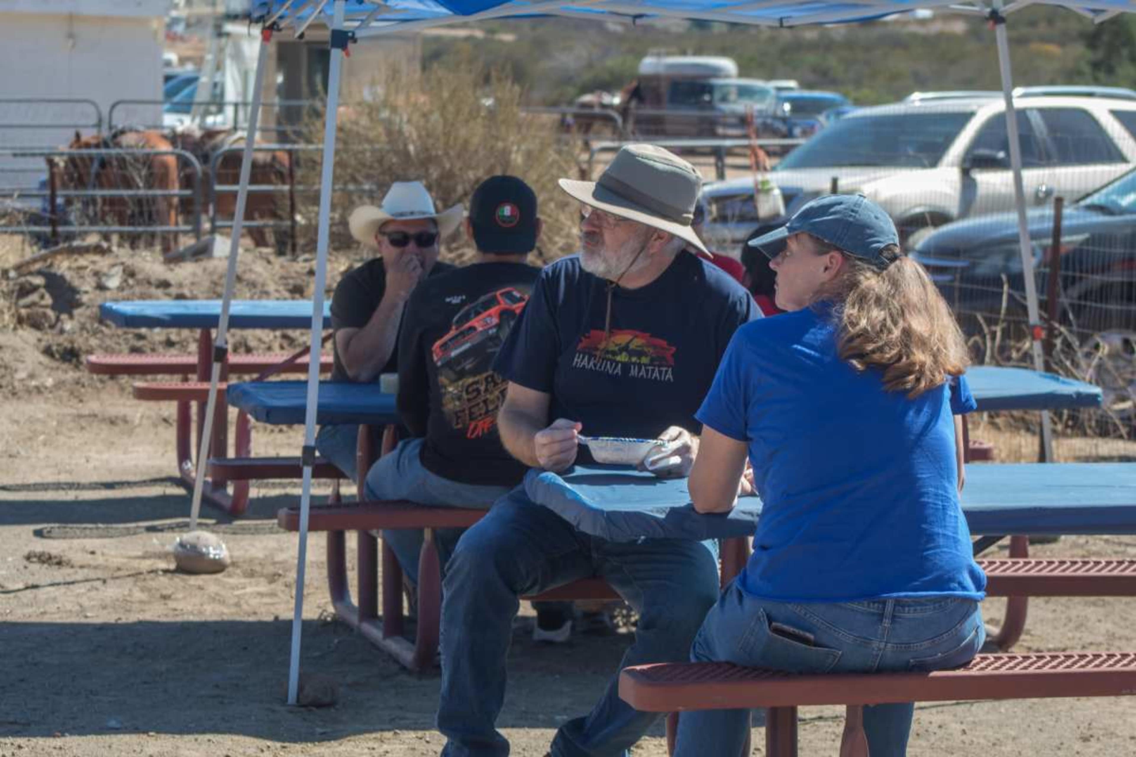 A group of people sits at blue picnic tables under a canopy in a rural outdoor setting.