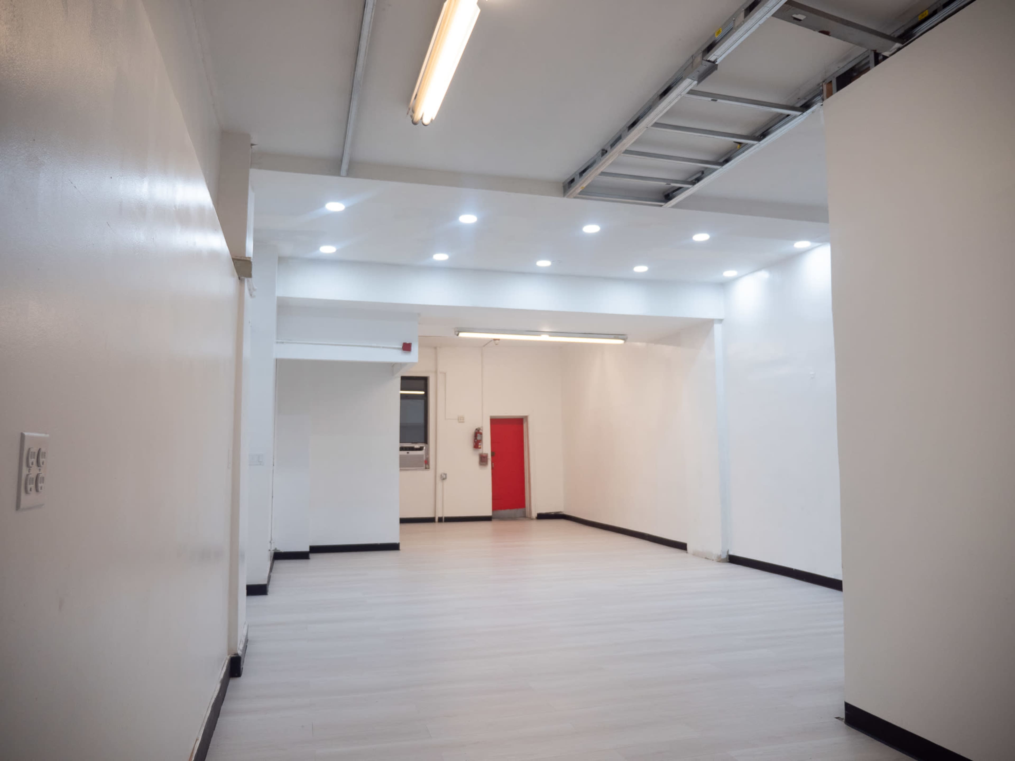 A large, empty room with white walls, a light-colored floor, and a red door at the far end.
