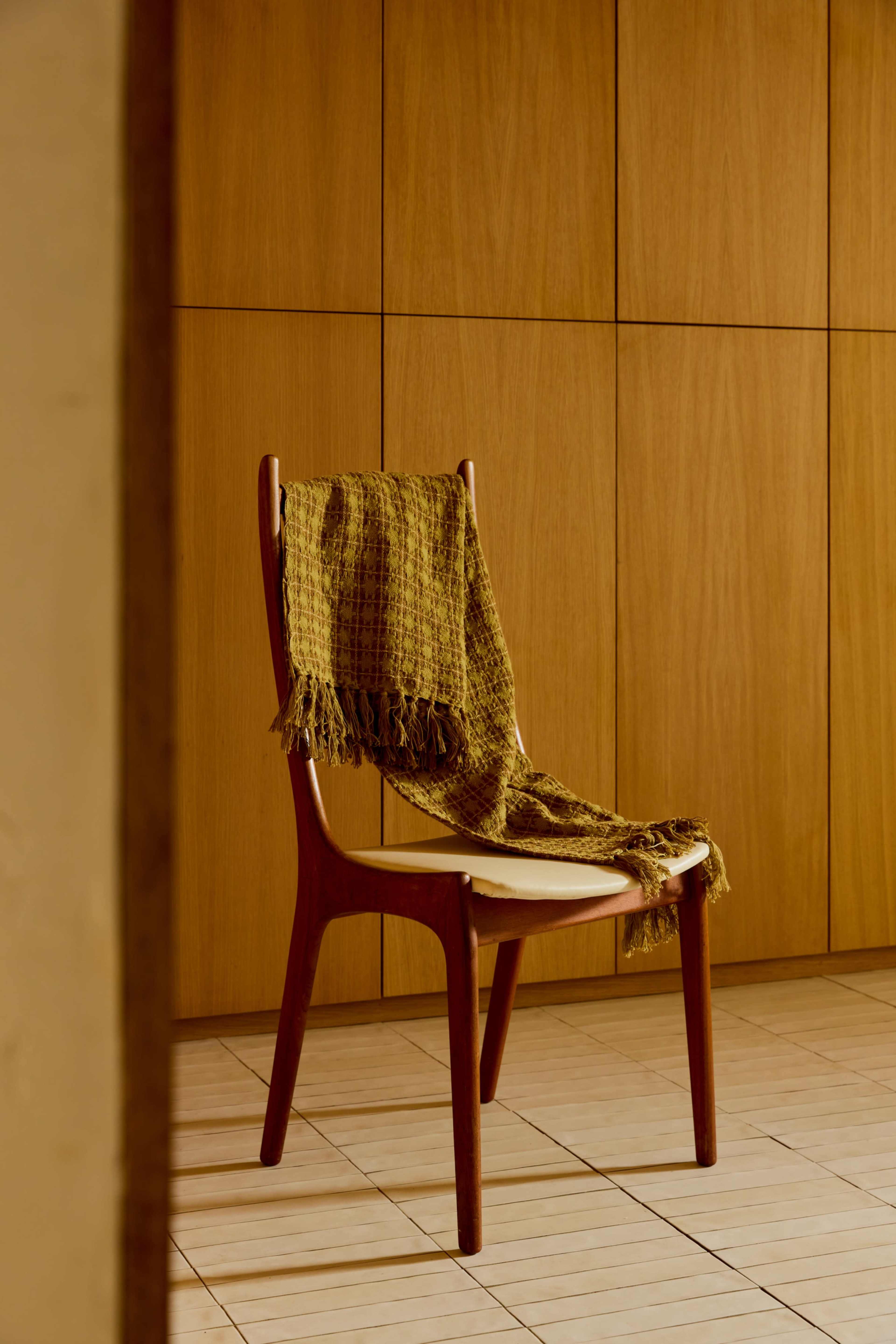 A wooden chair with a woven blanket draped over its back is positioned against a paneled wooden wall.