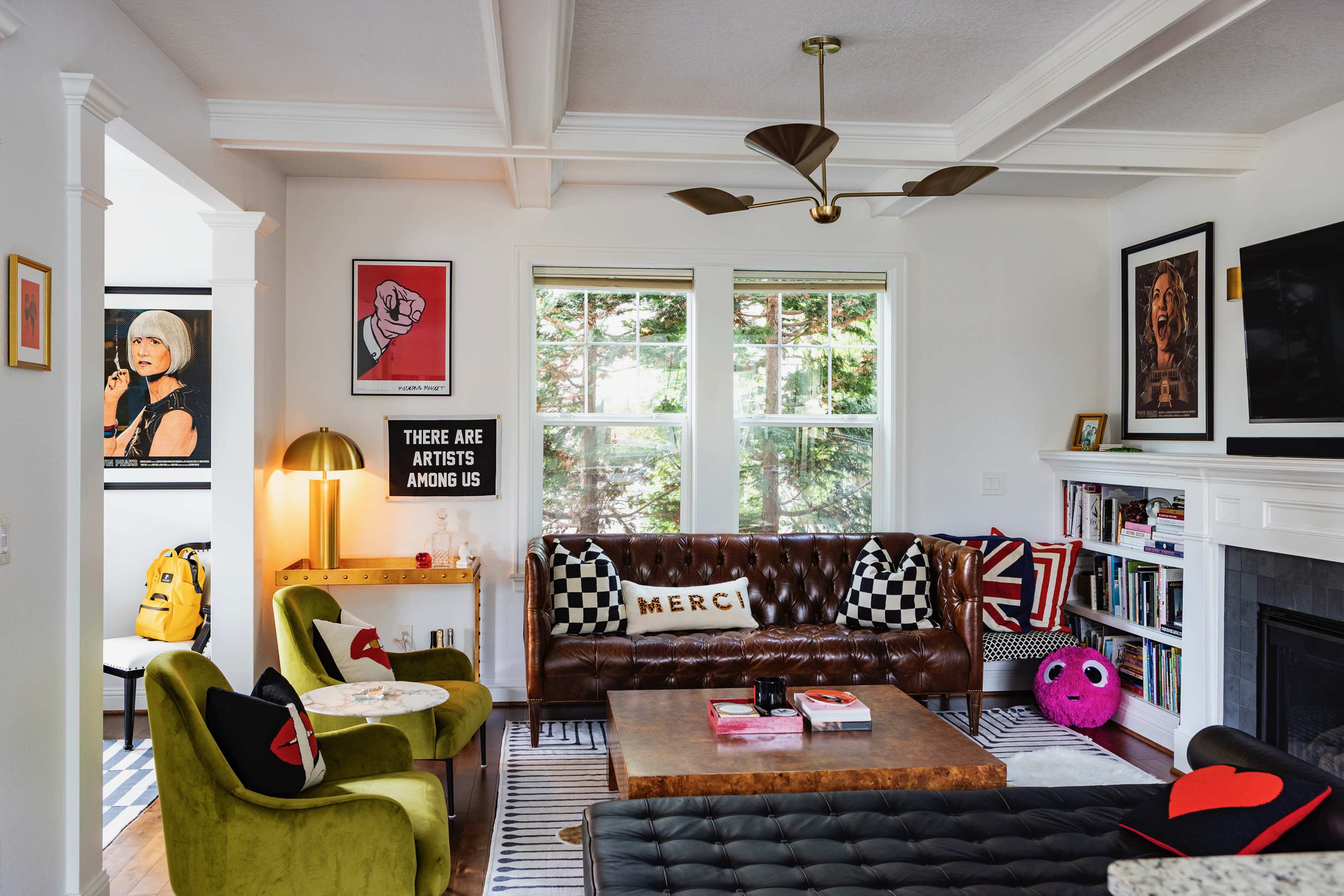 The living room features a mix of artwork on the walls, a brown leather sofa, green chairs, and a wooden coffee table, creating a cozy and eclectic atmosphere.