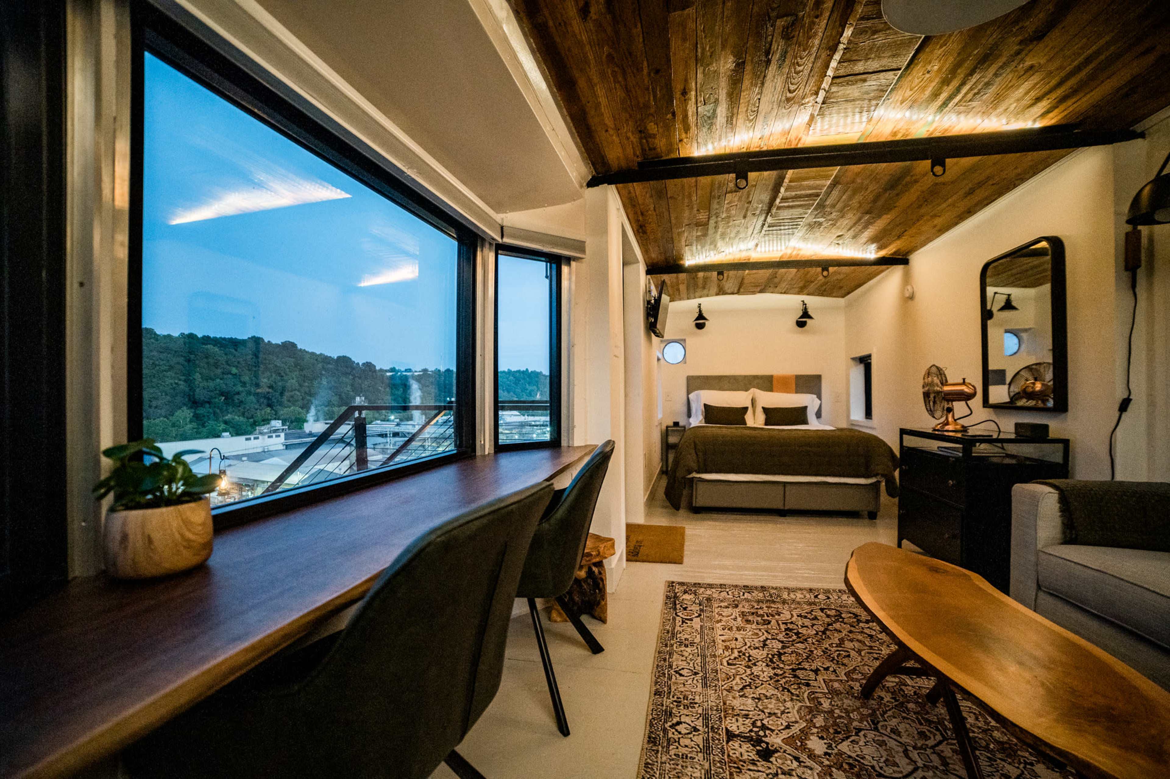 A modern interior of a small accommodation featuring a bedroom with a bed, two large windows, a small writing desk, a seating area, and wooden accents on the ceiling.
