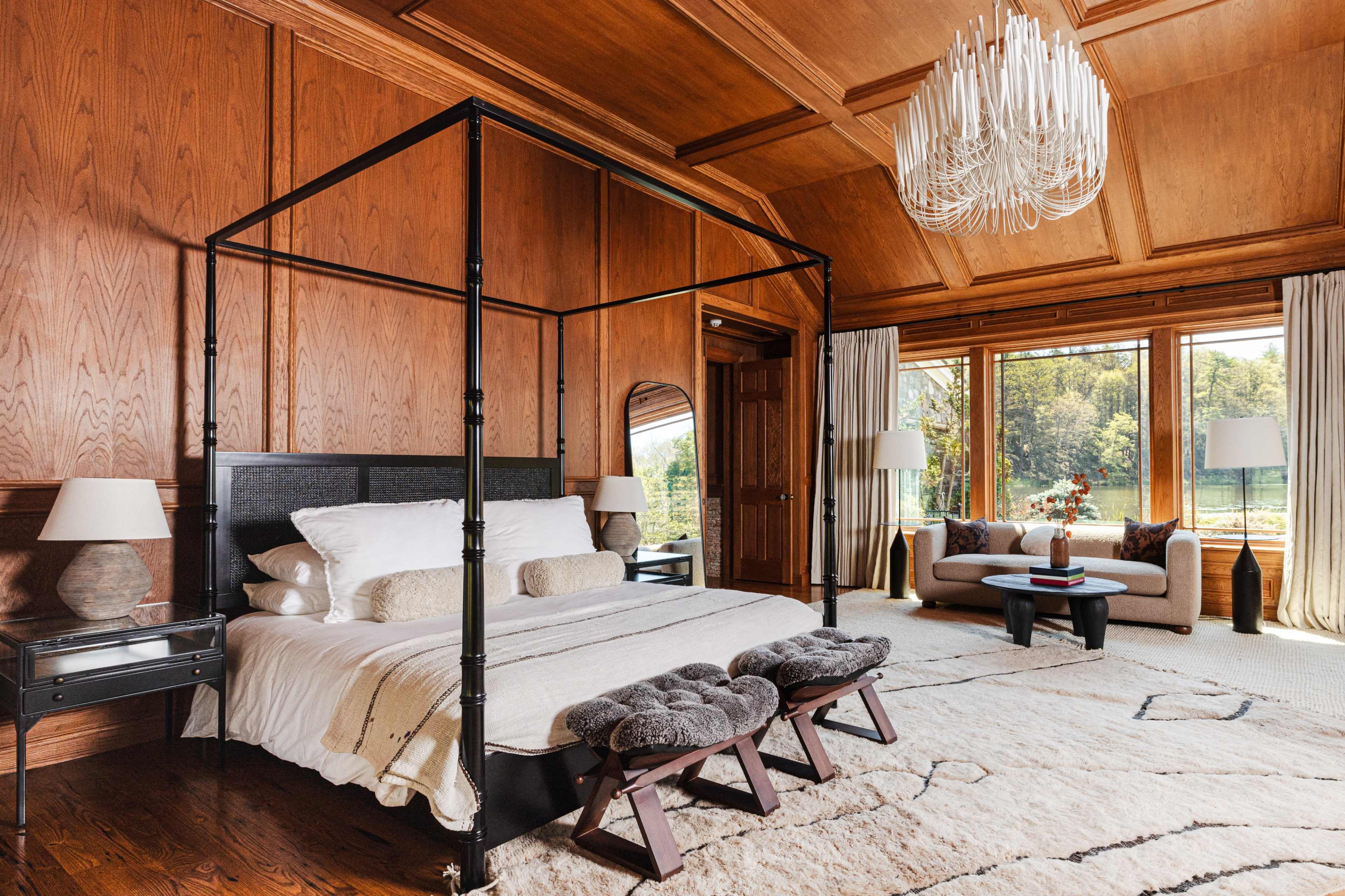 A spacious bedroom features a four-poster bed, two upholstered stools, and large windows with a view of the outdoors.