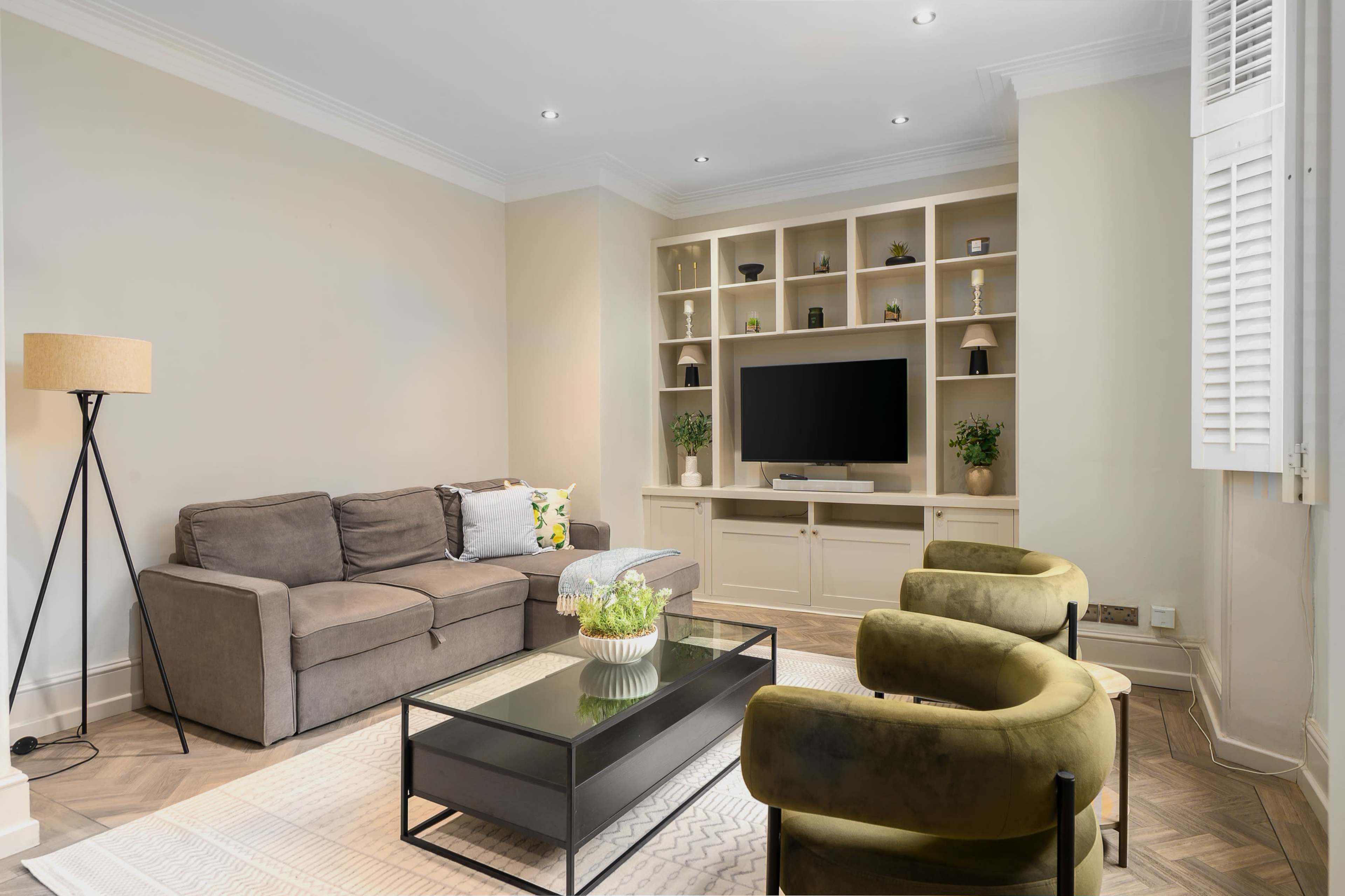 The image shows a modern living room with a gray sofa, two green armchairs, a glass coffee table, and a wall-mounted television surrounded by shelves.