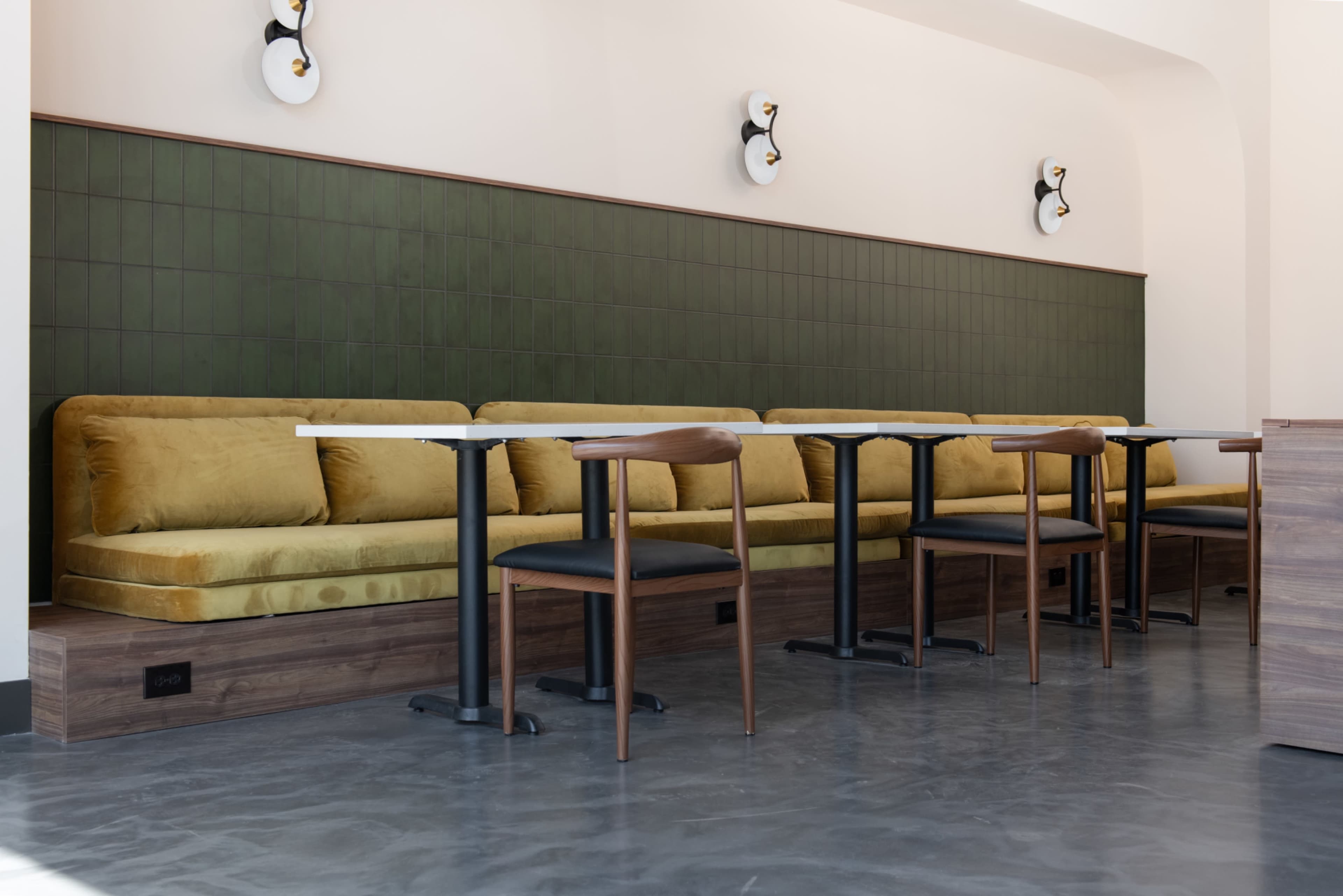 The image shows a modern café interior featuring a long, green-tiled wall with upholstered seating and a row of wooden tables and chairs.