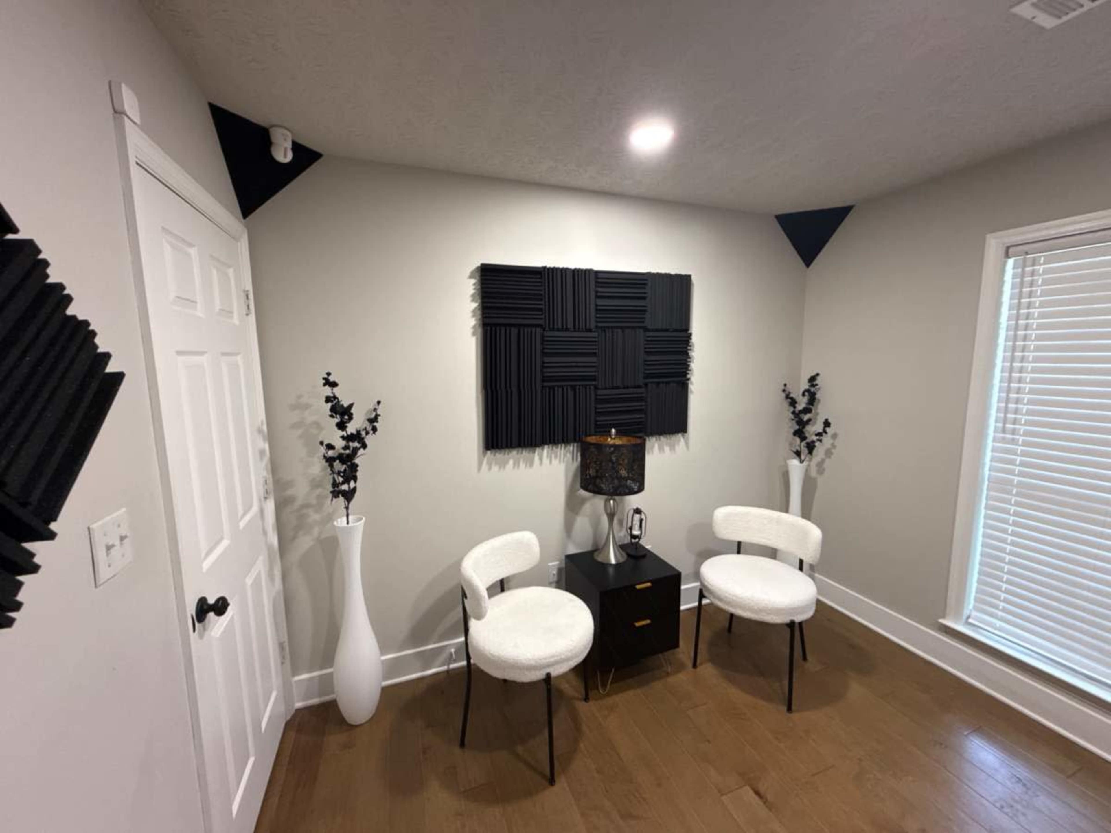 The image shows a small room with two white chairs, a black nightstand, and sound-absorbing panels mounted on the wall, along with decorative vases.