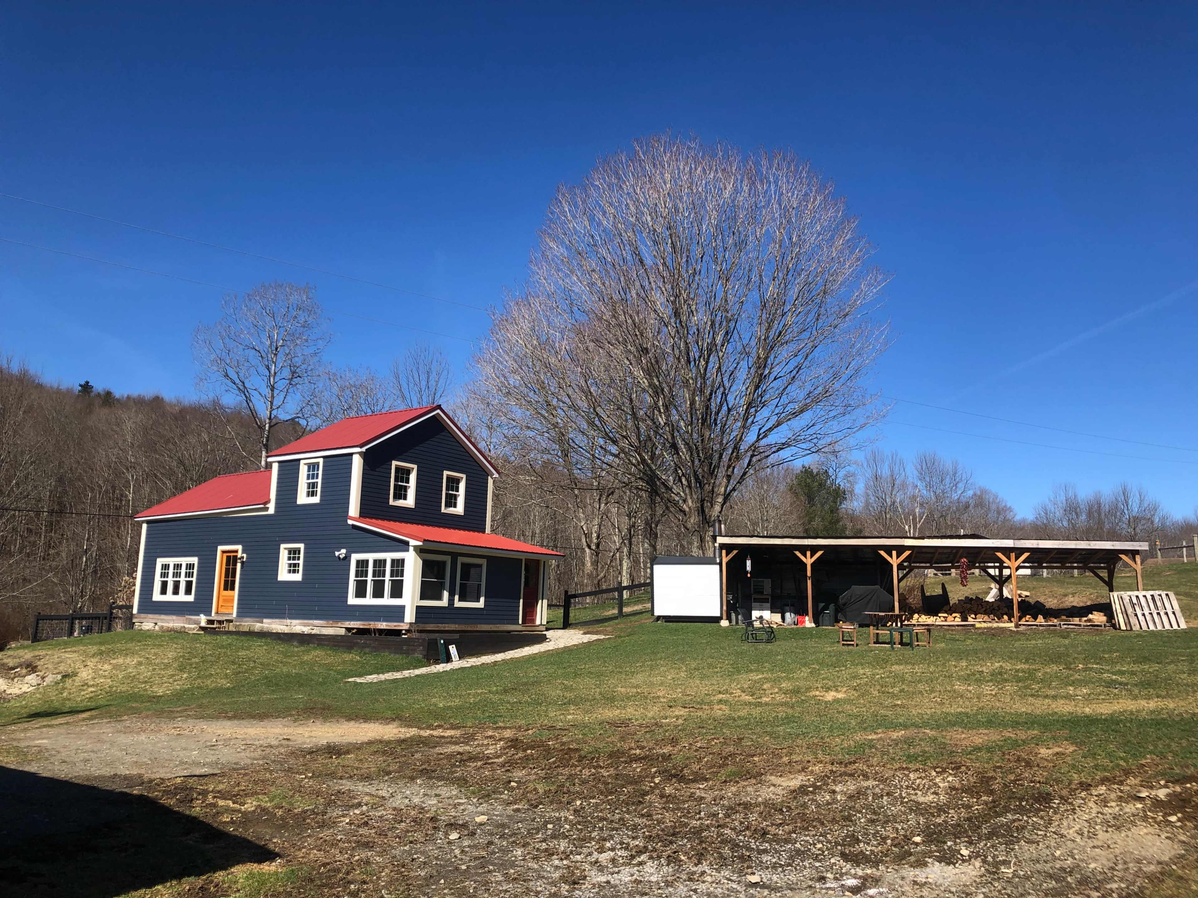 A two-story house with a red roof and blue siding is situated next to a large tree and a covered structure in an open field.