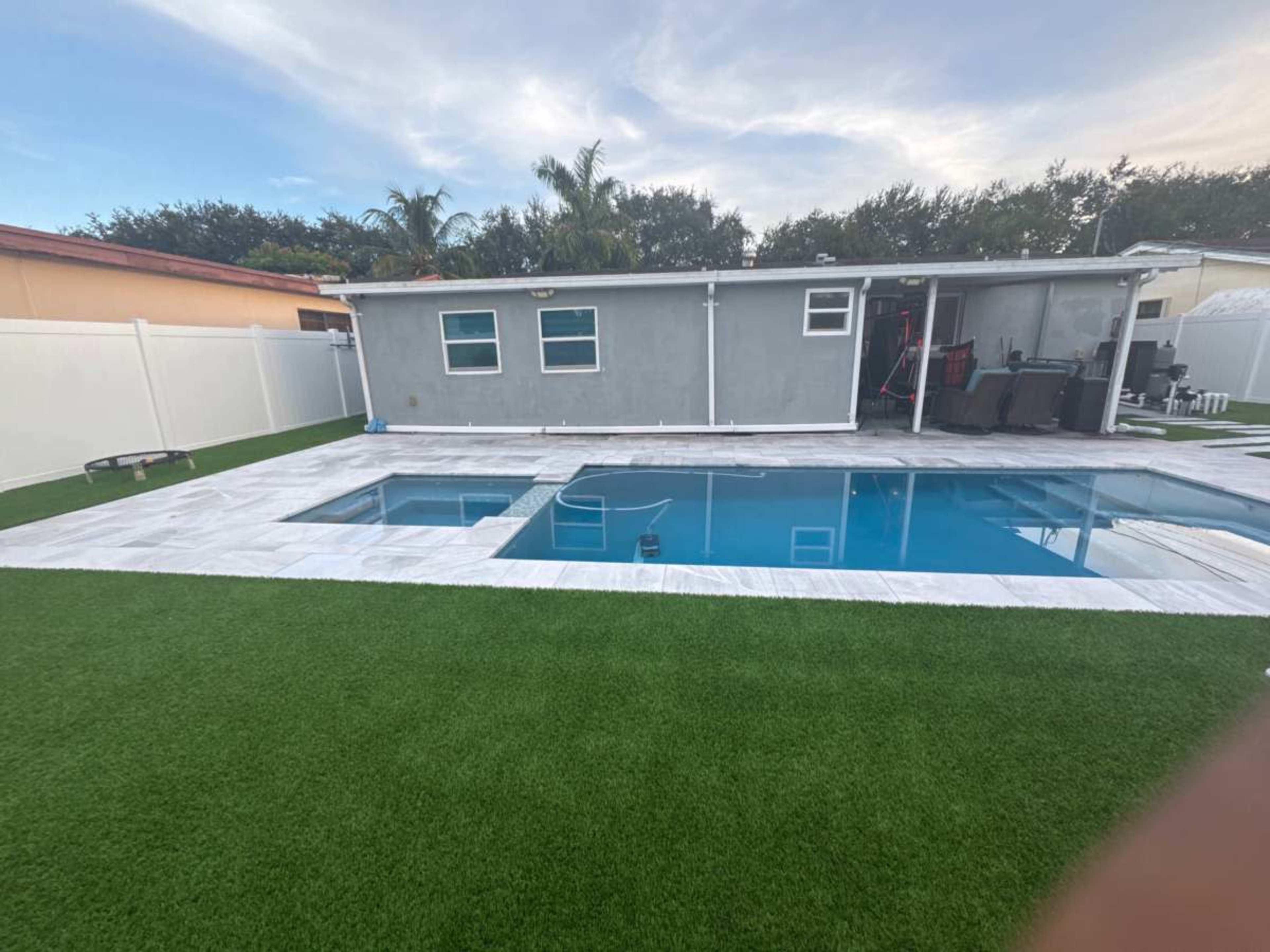 The image shows a backyard with a swimming pool surrounded by a stone patio and artificial grass, with a gray shed or structure in the background.