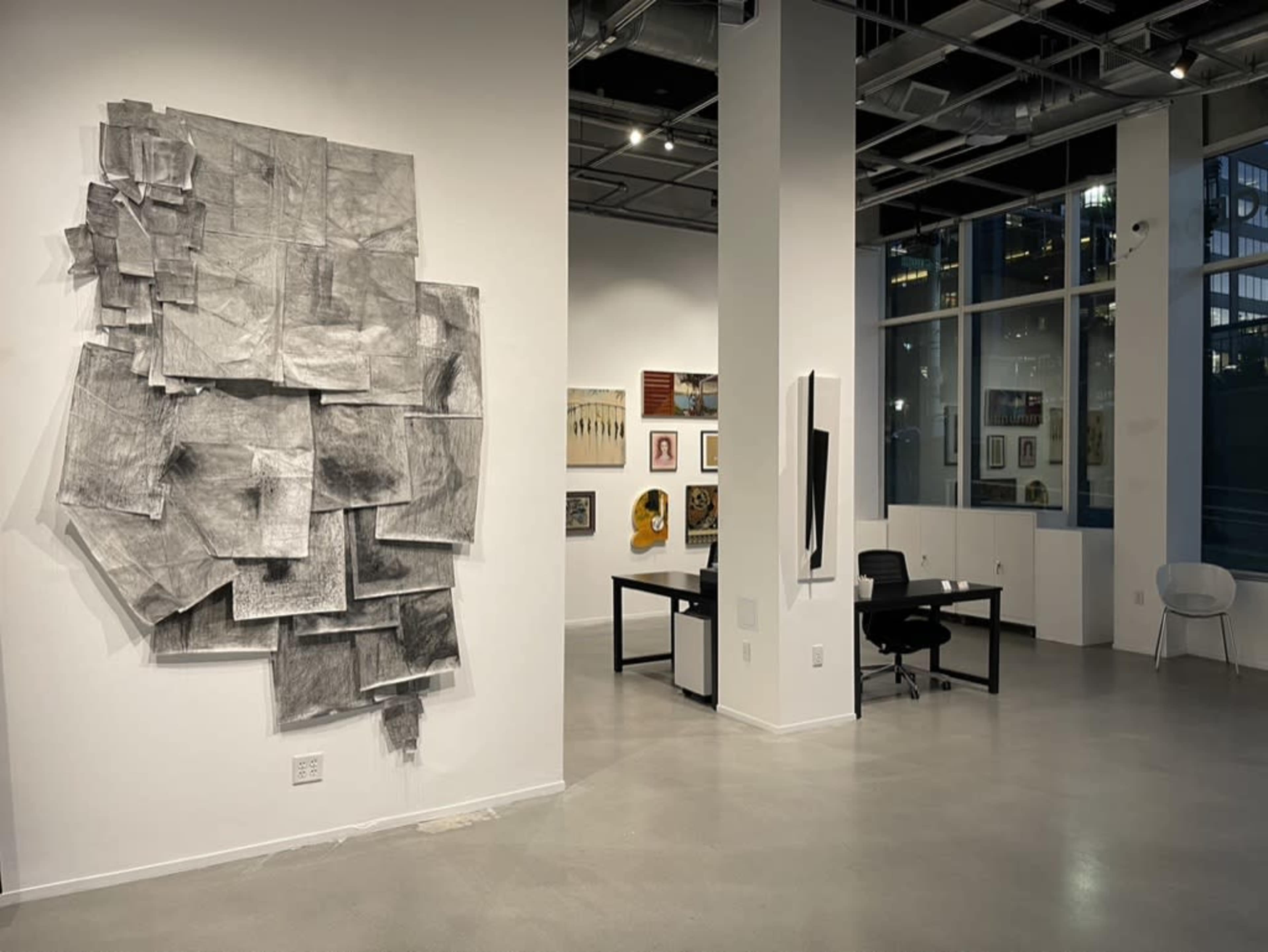 The image shows an art gallery featuring a large charcoal artwork on one wall and various framed artworks displayed on another wall, with a desk and chair visible in the foreground.