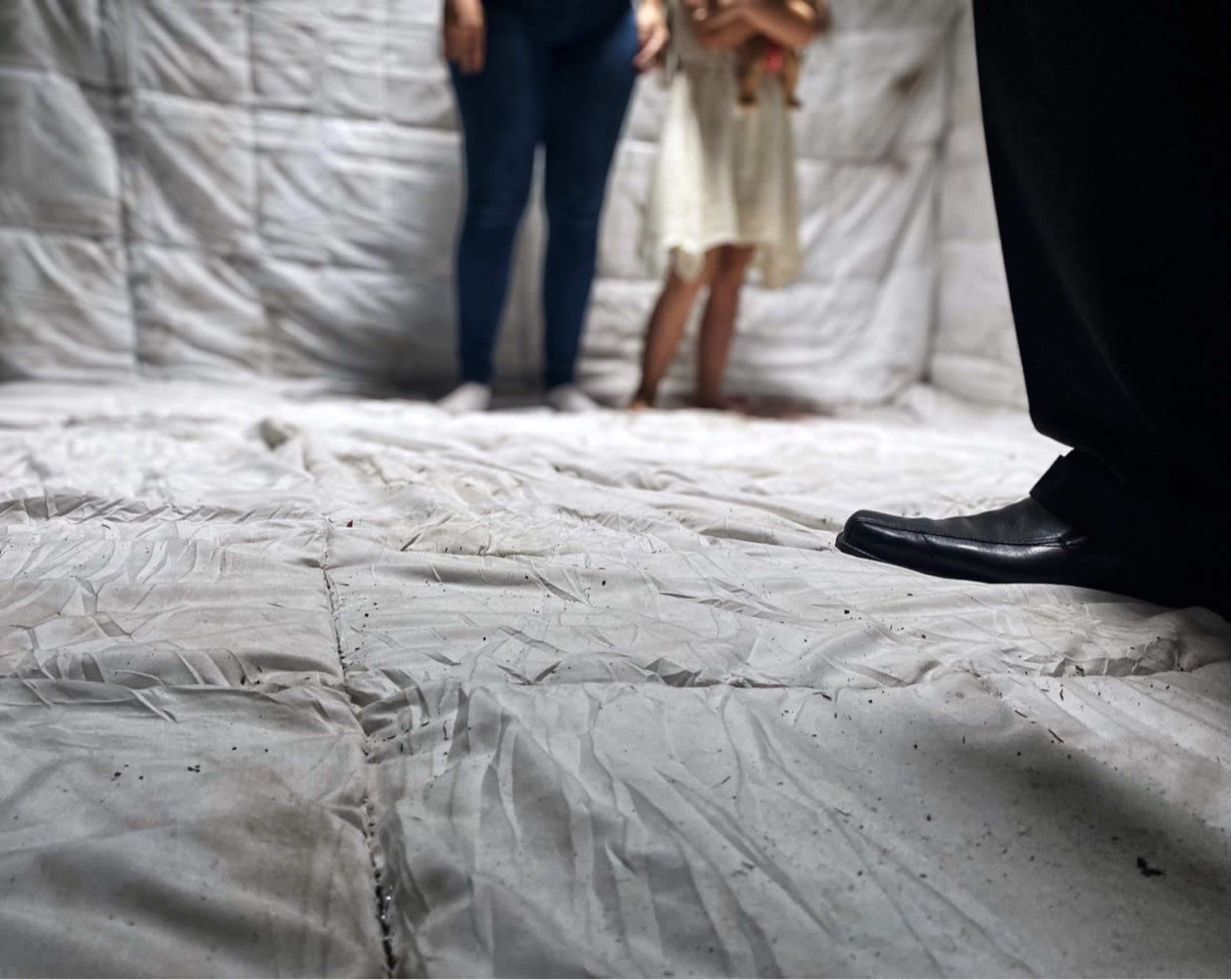 Two figures stand on a wrinkled, white surface in a dimly lit space with a textured backdrop.