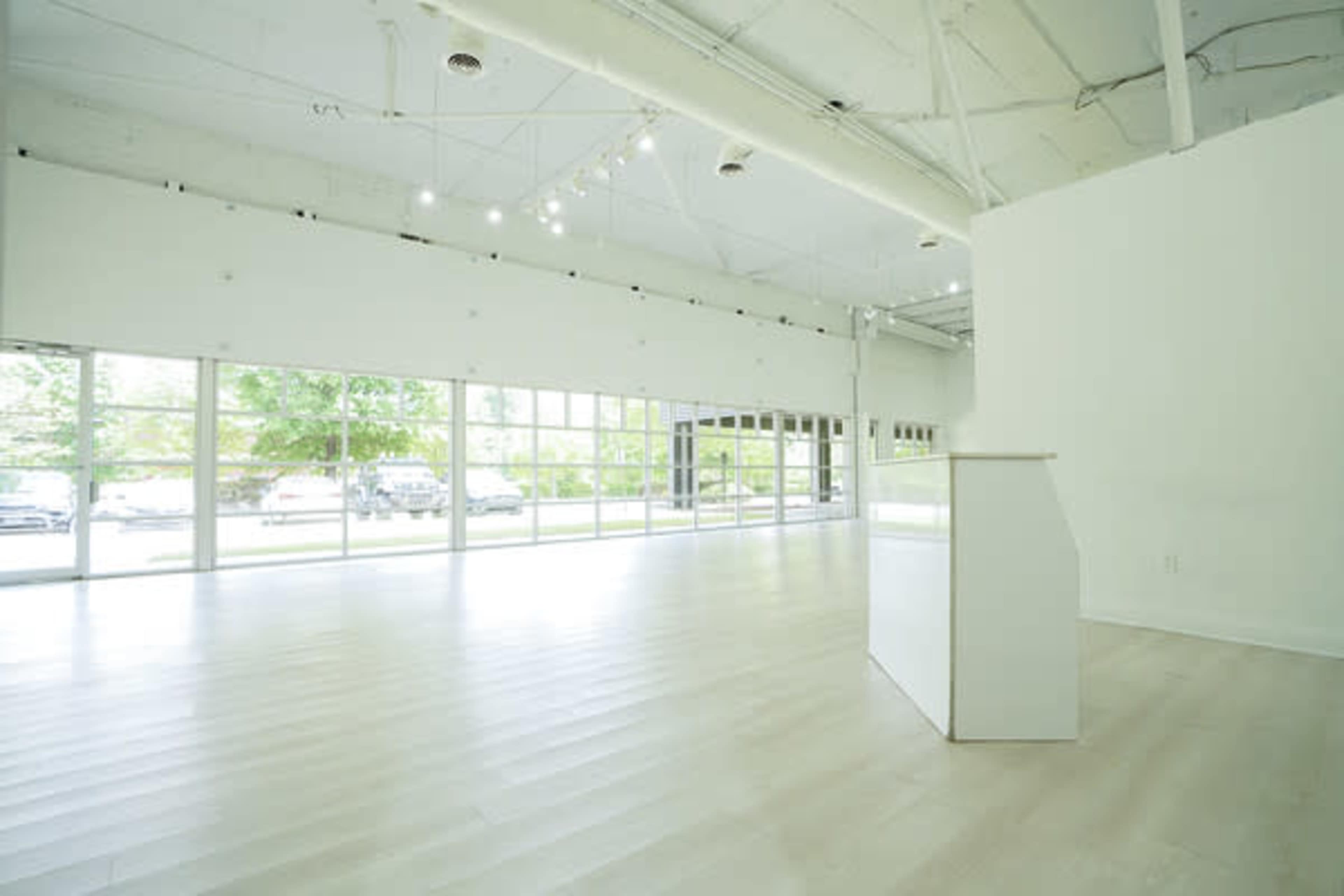 The image shows a spacious, well-lit interior of a modern room with large windows and minimal furnishings.