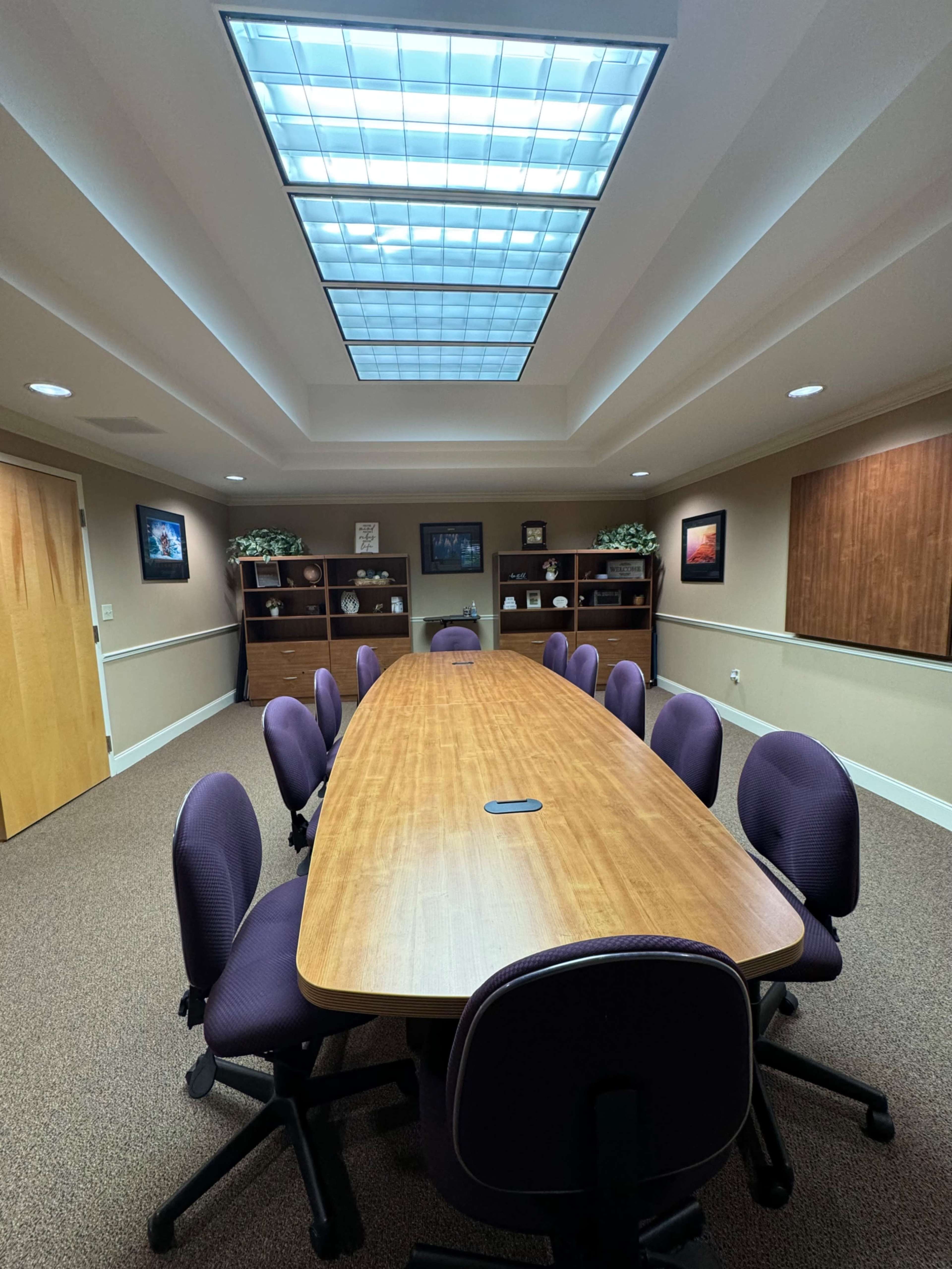 A long wooden conference table surrounded by purple chairs is illuminated by natural light from a skylight overhead in a professional meeting room.