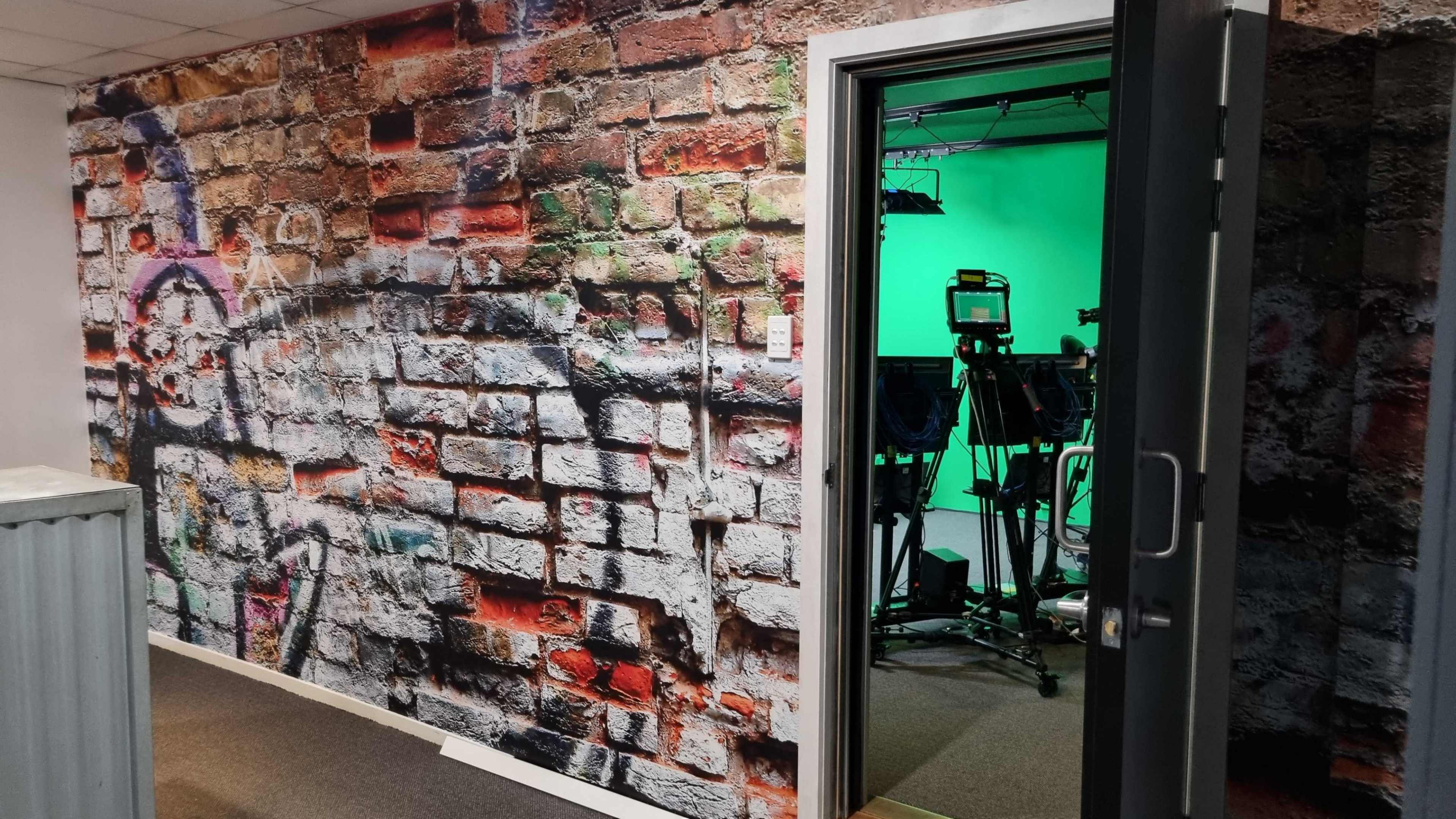 The image shows a room with a brick wall mural on one side and an open door leading into a space with green screen equipment inside.
