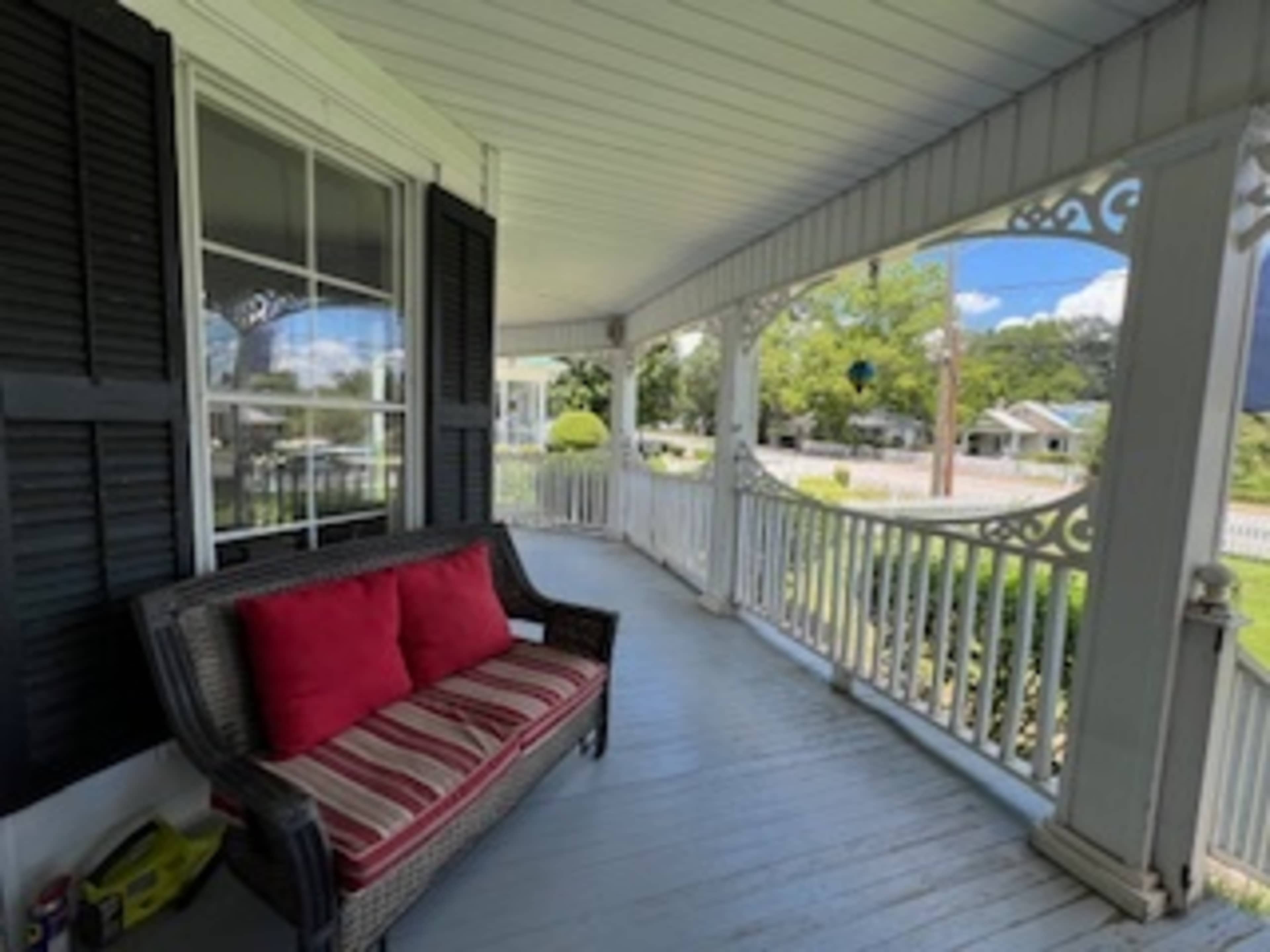 A covered porch features a wicker sofa with red cushions, overlooking a neighborhood lined with trees and houses.