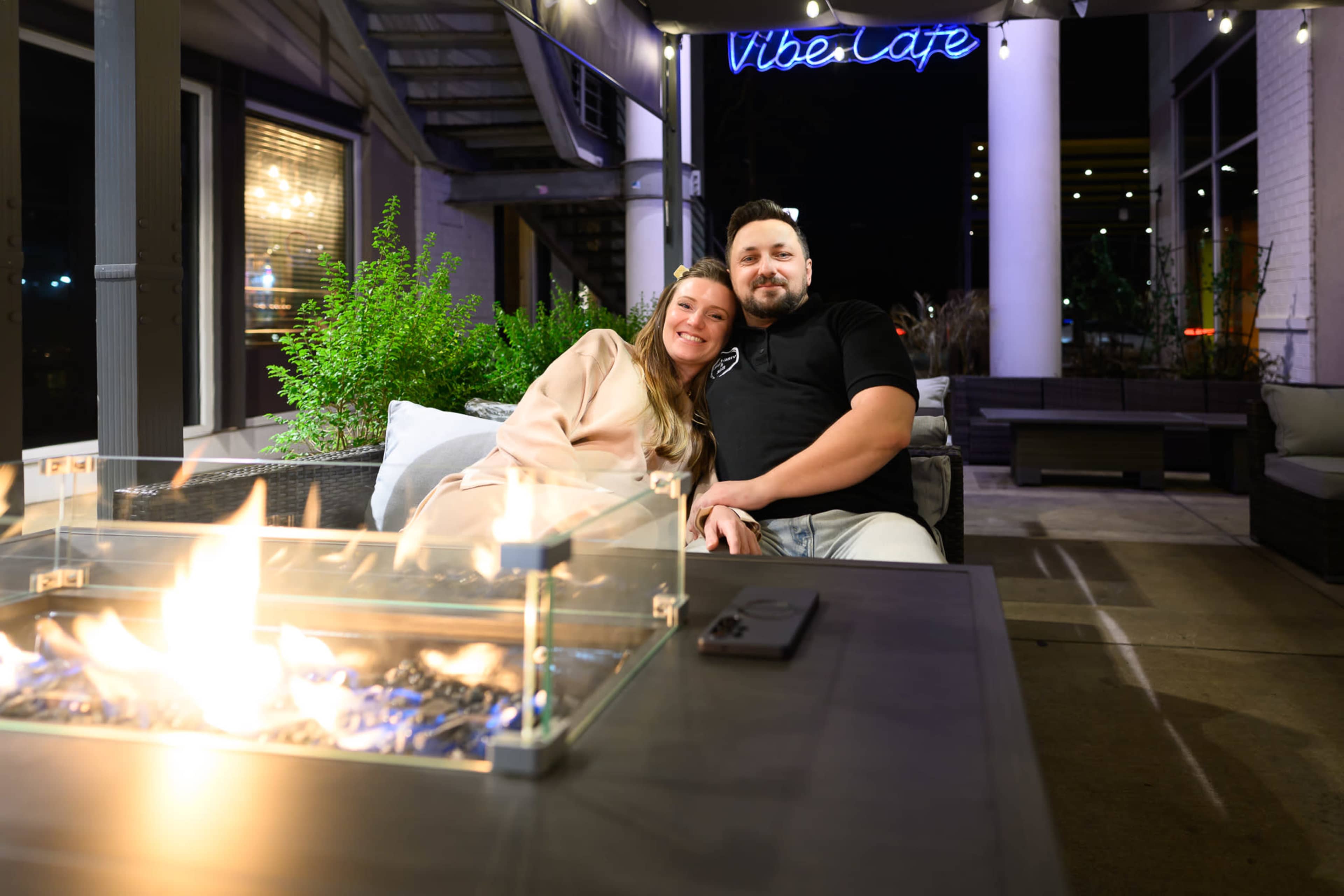 A couple sits together on a sofa near a fire pit at a café, with a glowing sign that reads "Vibe Café" in the background.