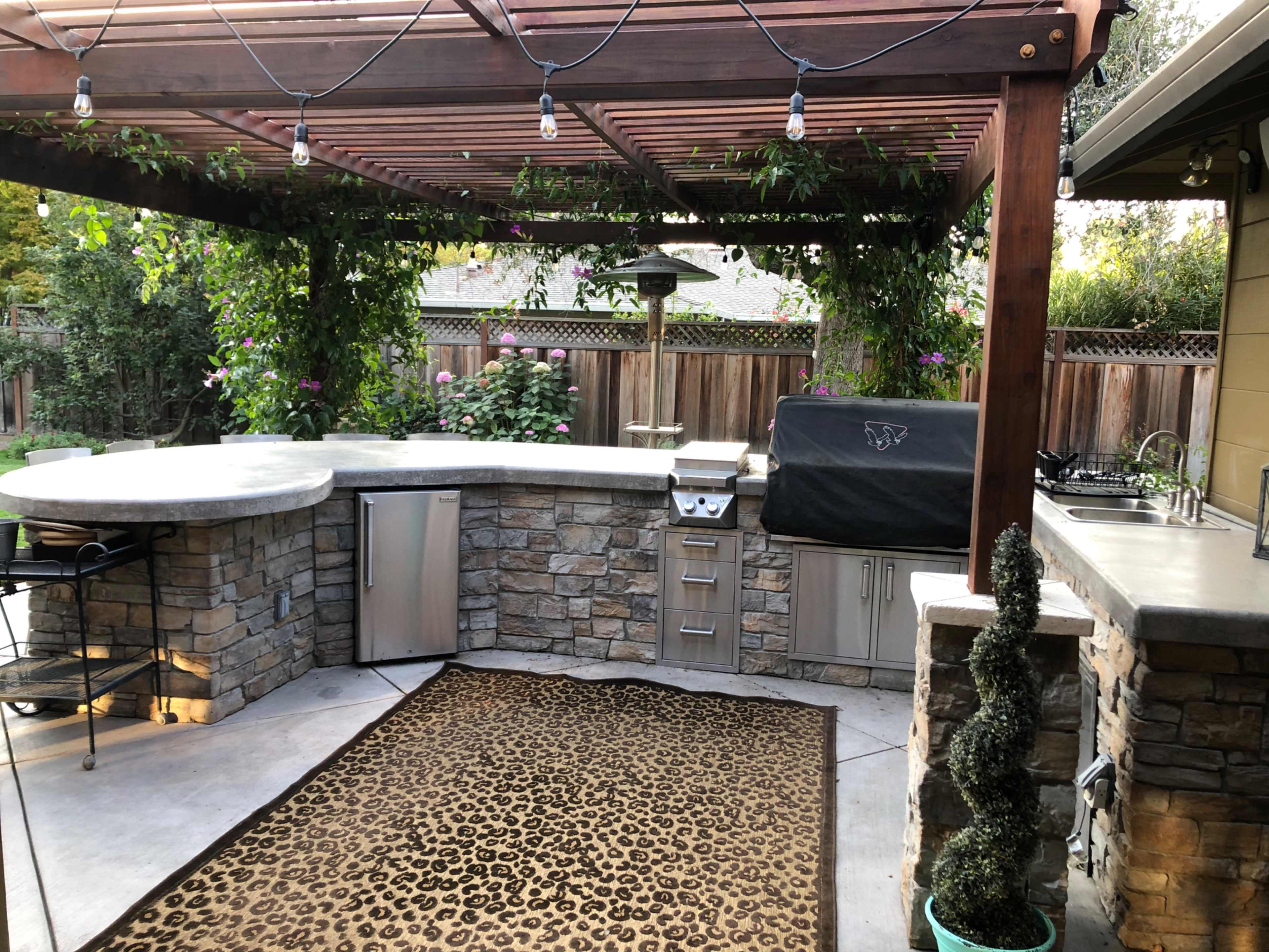 An outdoor kitchen area with a stone countertop, a grill, a refrigerator, and a pergola with hanging lights and greenery.
