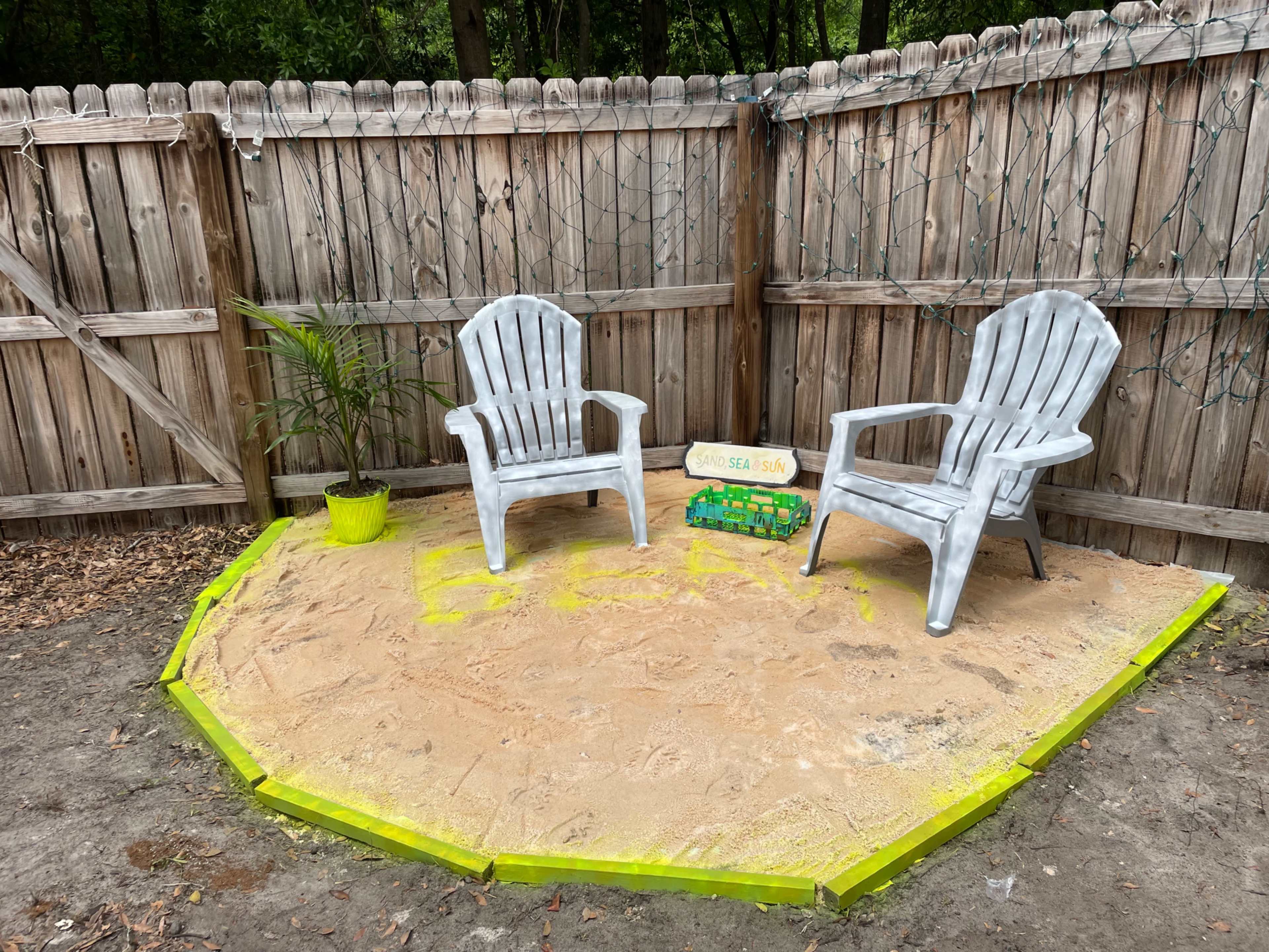 Two white plastic chairs sit on a sandy patch surrounded by a wooden fence, with a potted plant and a small sign that reads "Good Vibes Only."