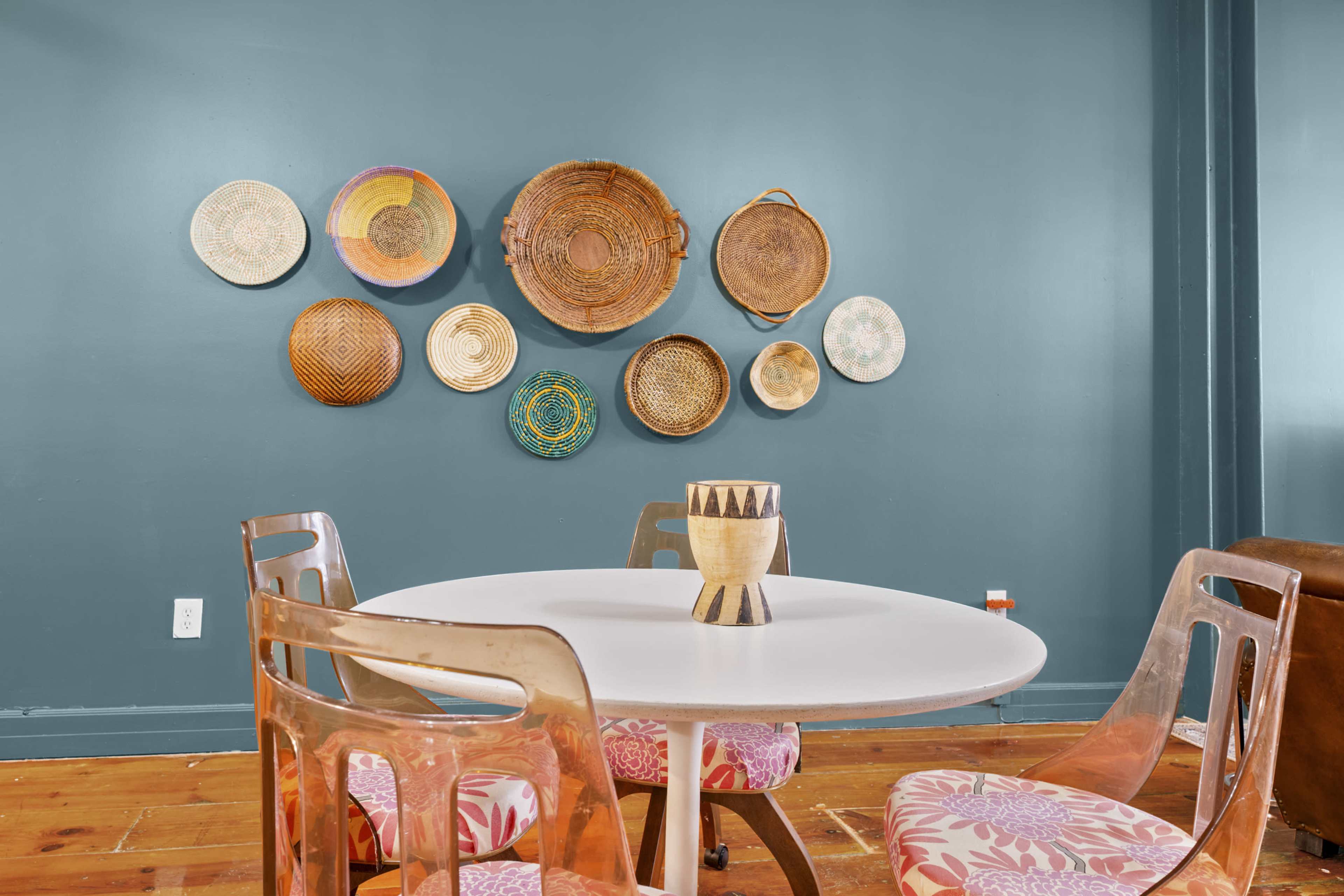 A round table with clear chairs is positioned in front of a wall adorned with various woven baskets in different sizes and designs.