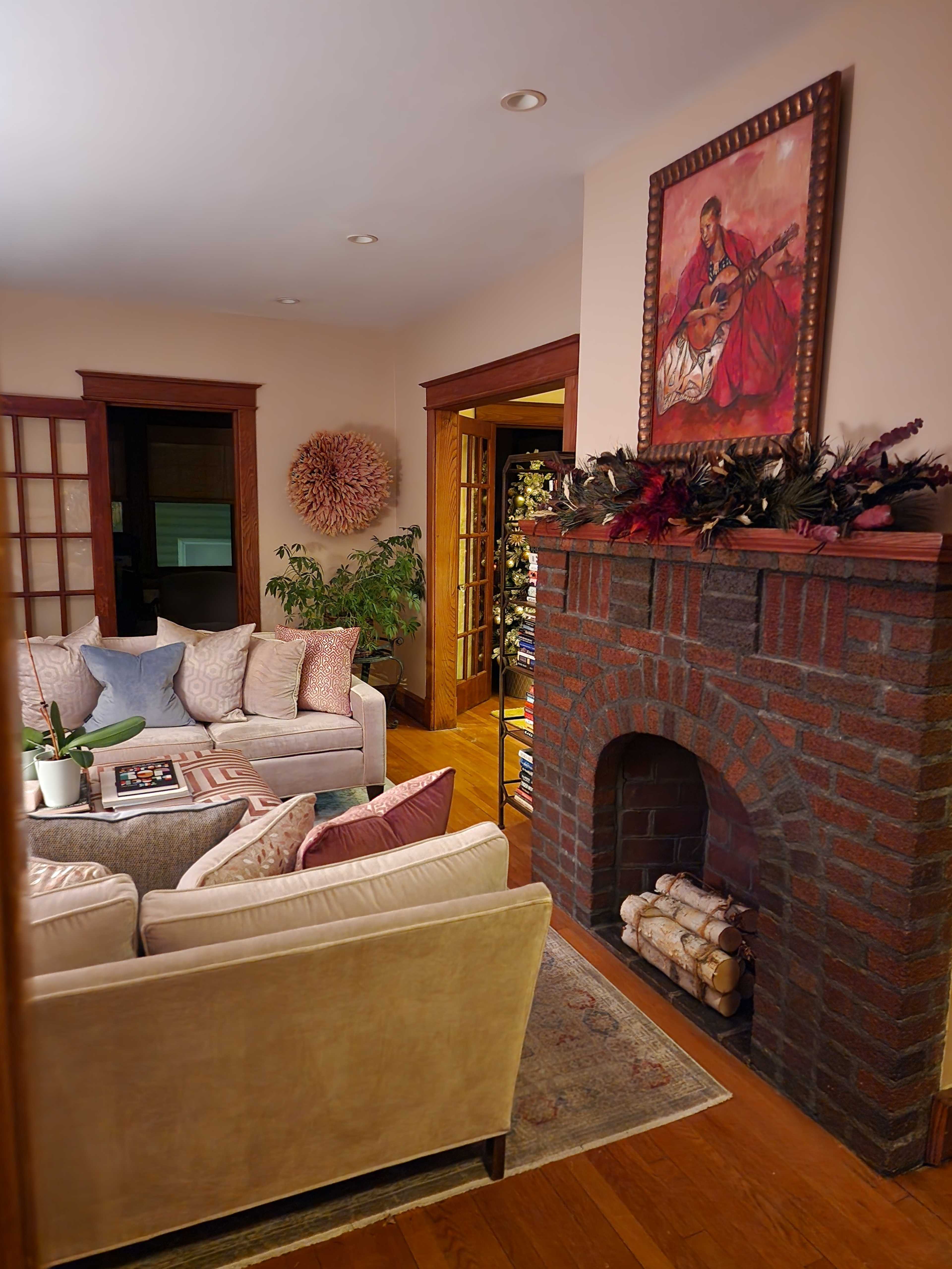A cozy living room features a sectional sofa, a vintage armchair, a brick fireplace with stacked logs, and a painting of a musician on the wall.