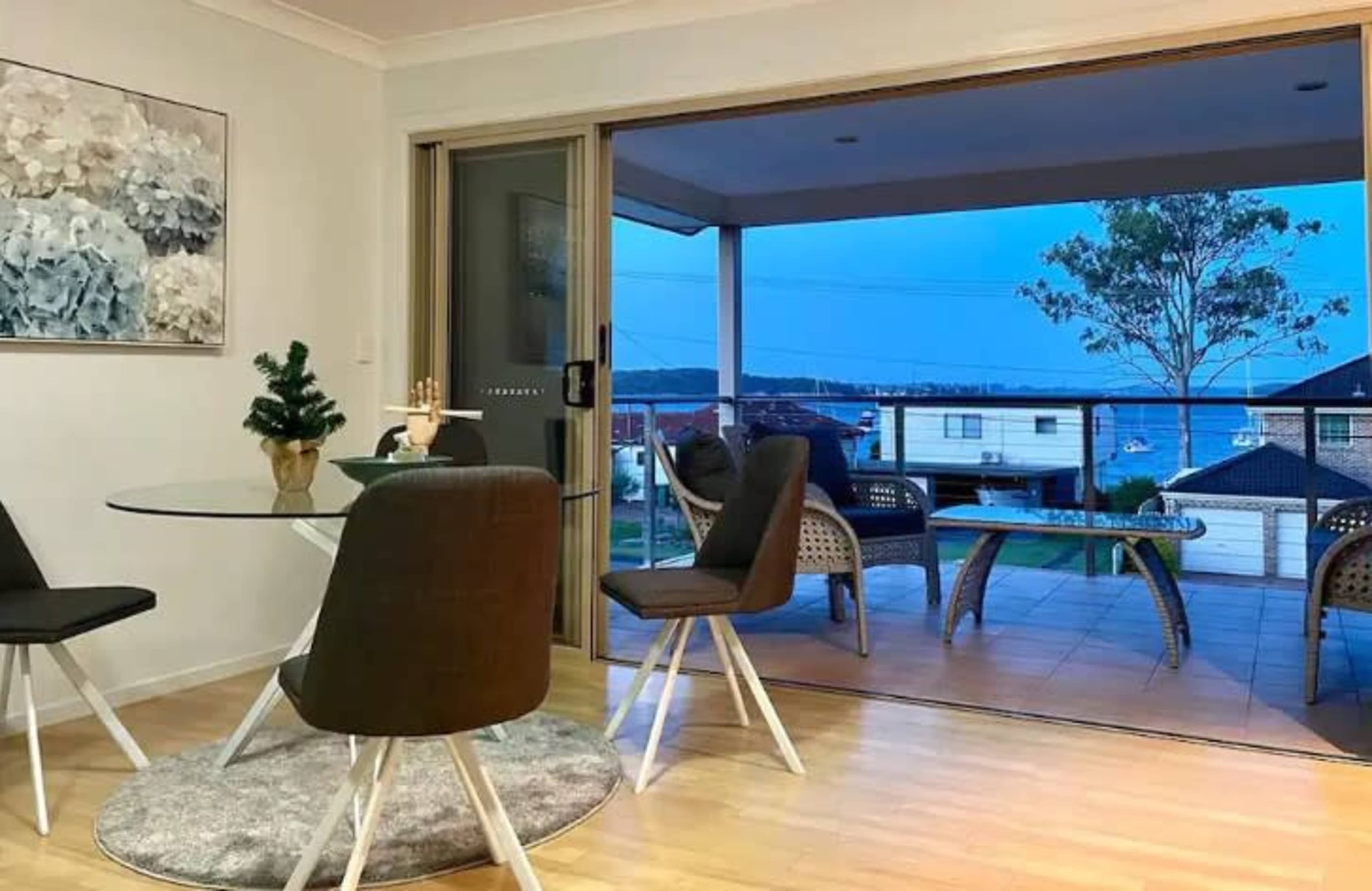 A dining area features a round glass table with four chairs and a sliding door that opens to a balcony overlooking a water view.