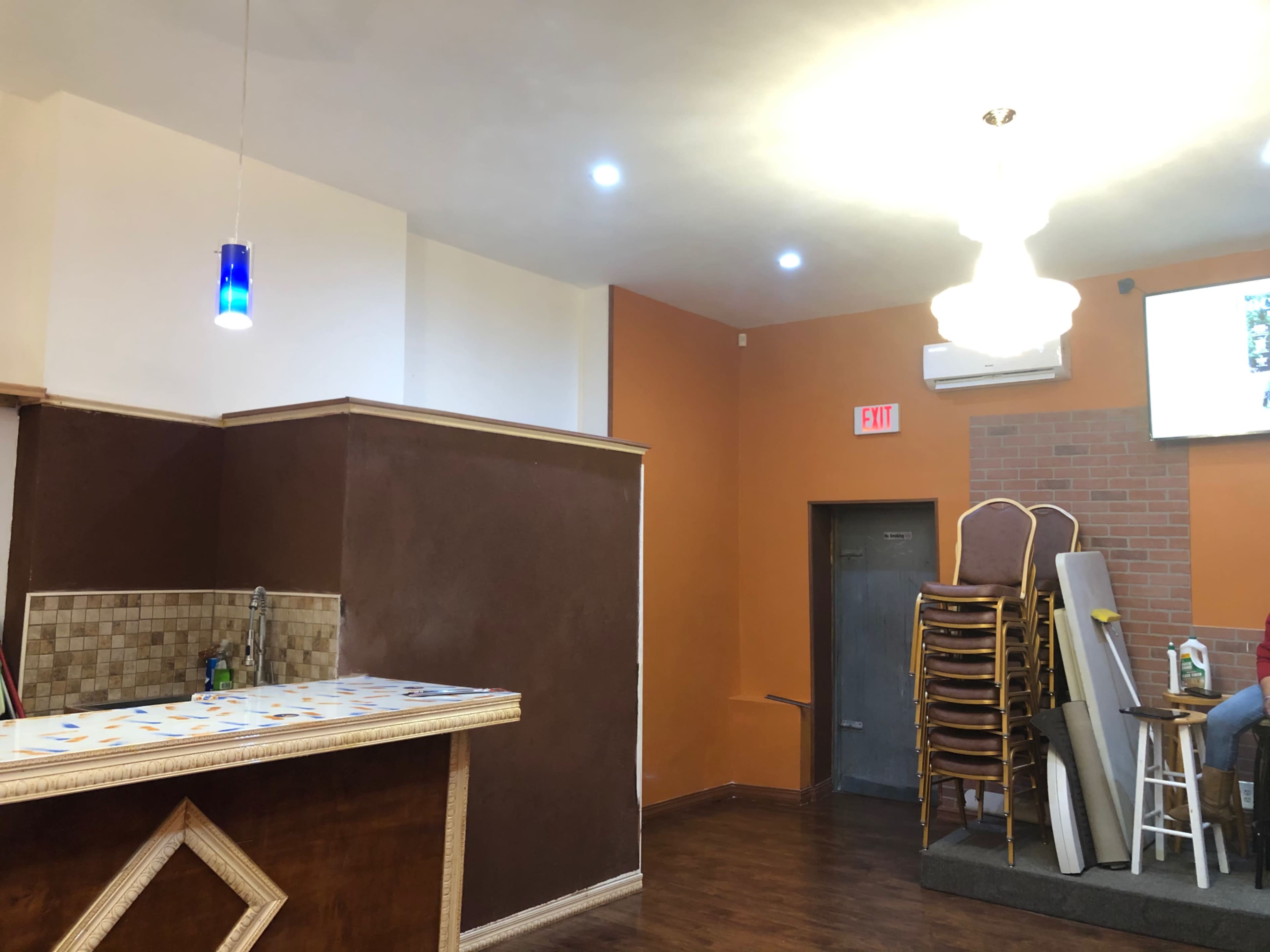 The interior of a restaurant features a bar with a patterned counter, orange walls, stacked chairs, and a decorative light fixture.