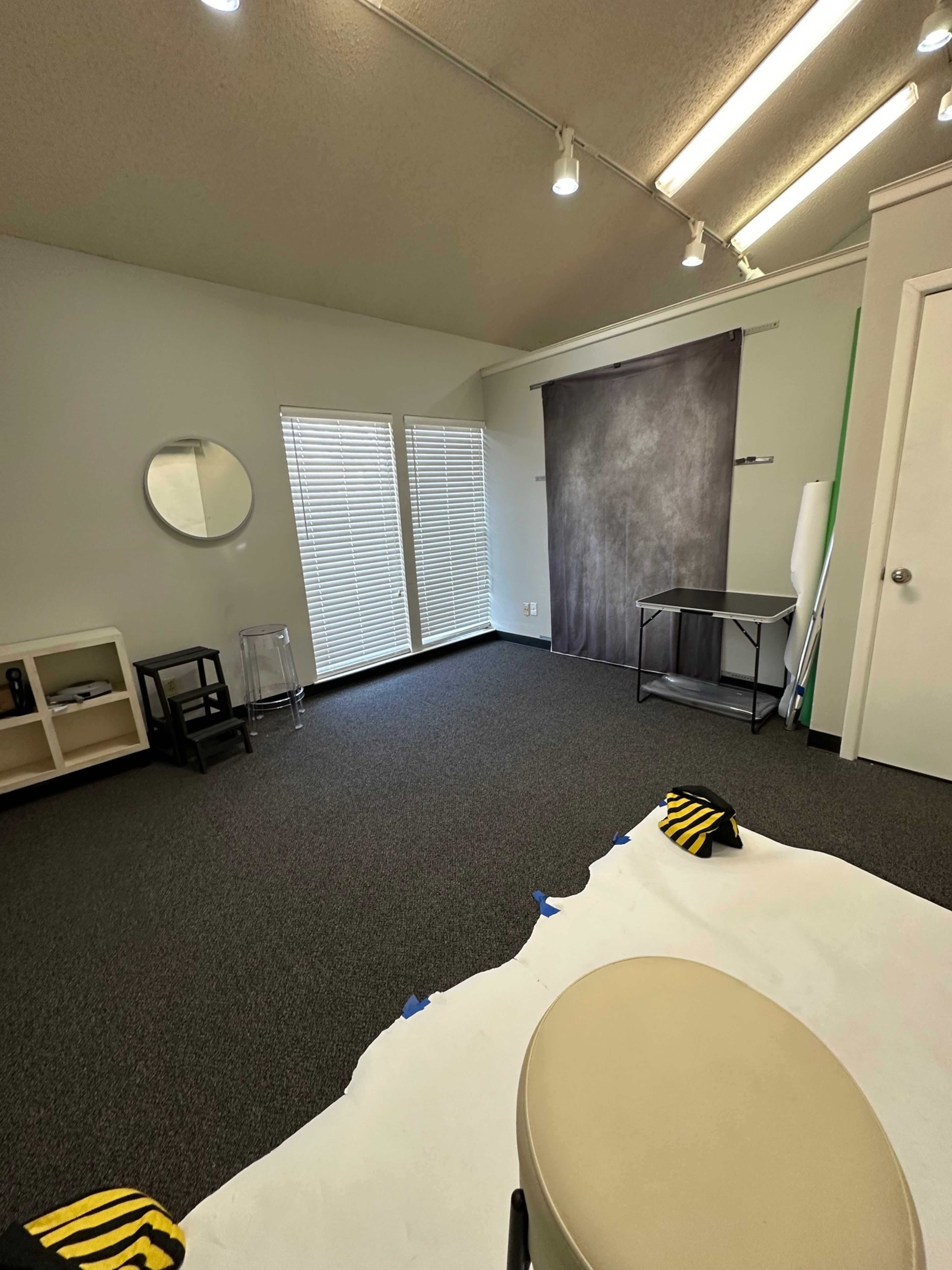 The image shows a spacious room with gray walls, a simple backdrop setup, and various furniture, including a stool and storage shelves.