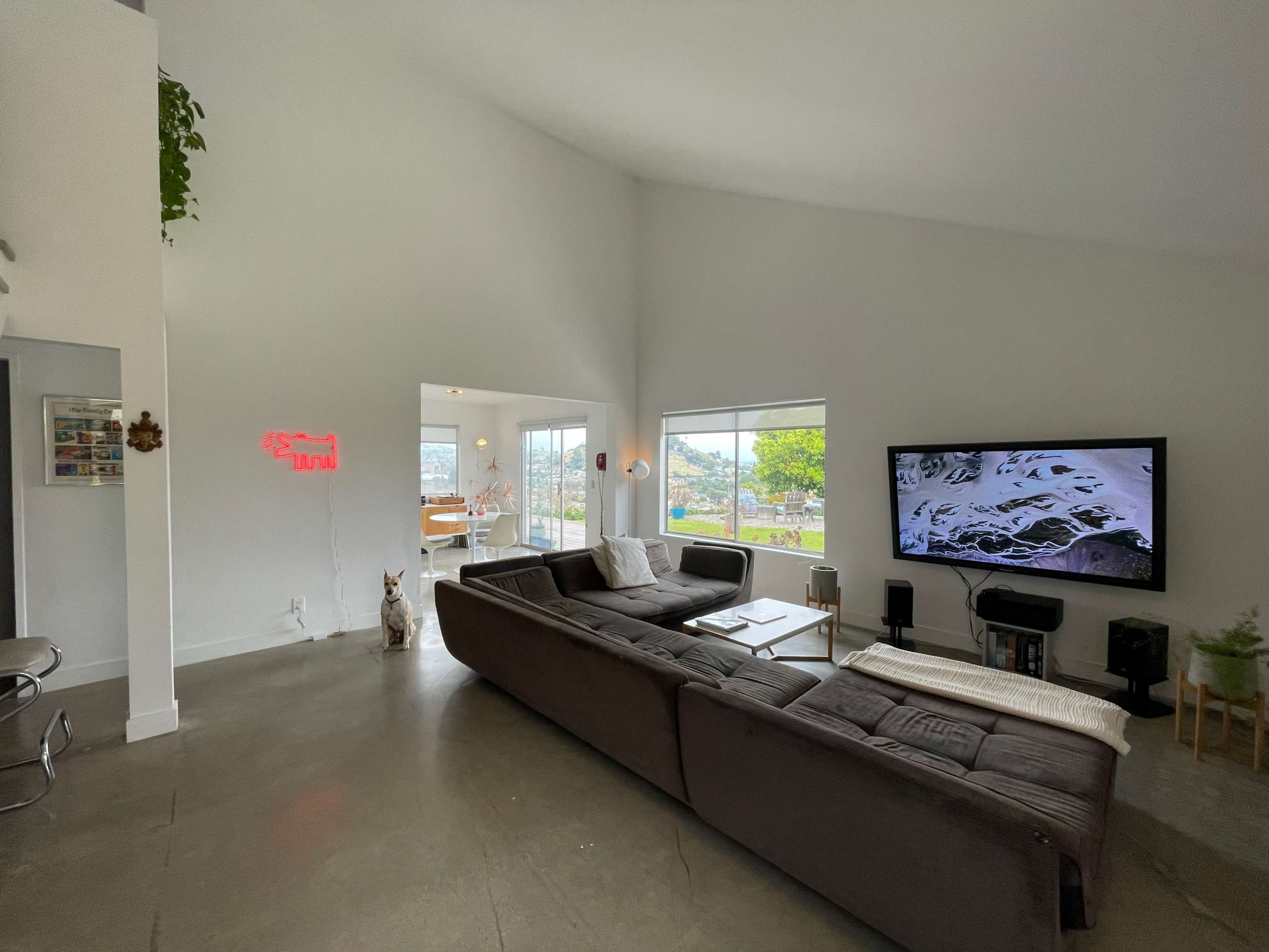 The image shows a modern living room with a large sectional sofa, a flat-screen TV on the wall, and large windows providing a view of the outdoors.