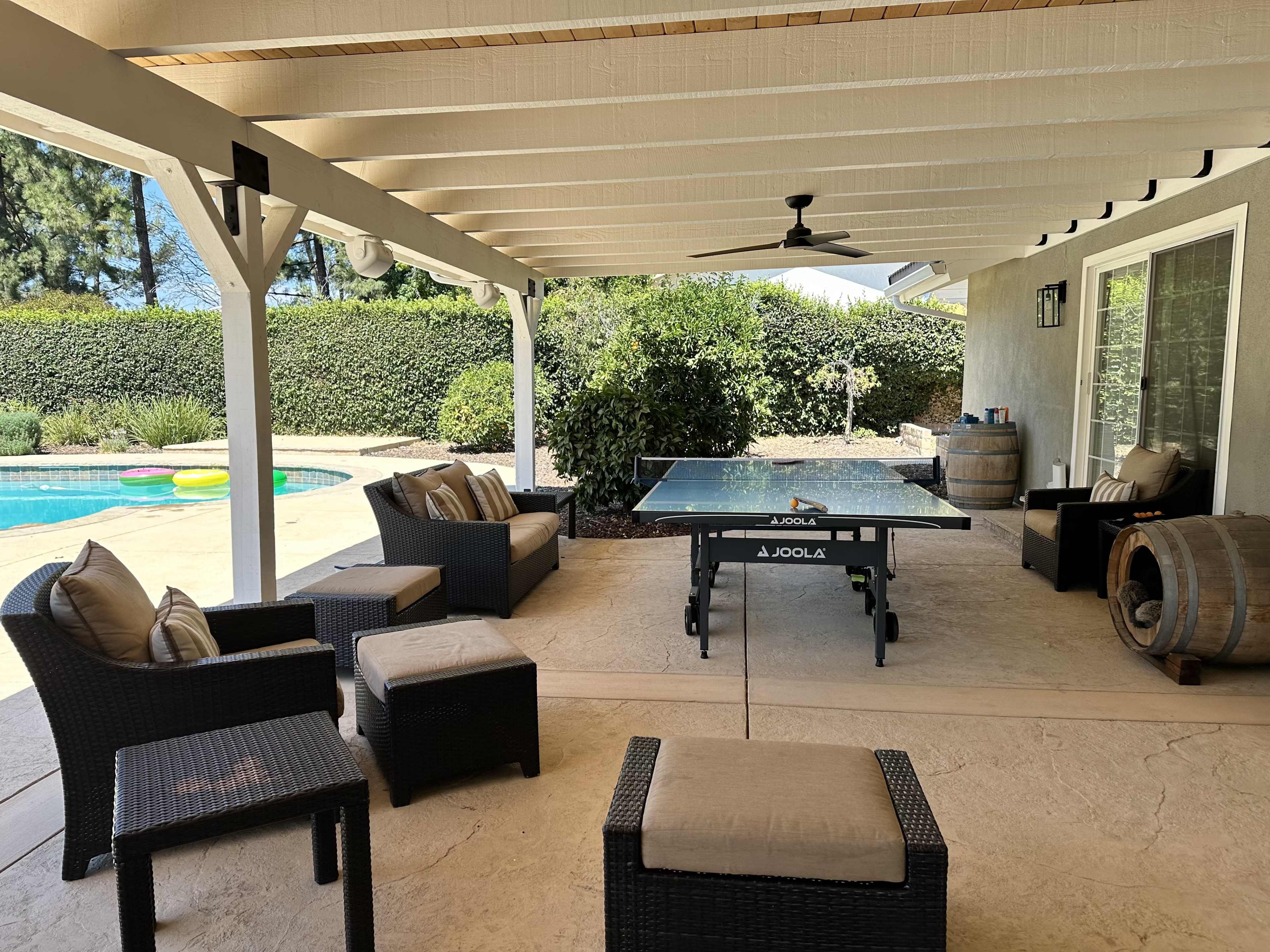 The image shows a covered outdoor patio area with a table tennis table, seating arrangements, and a view of a swimming pool in the background.