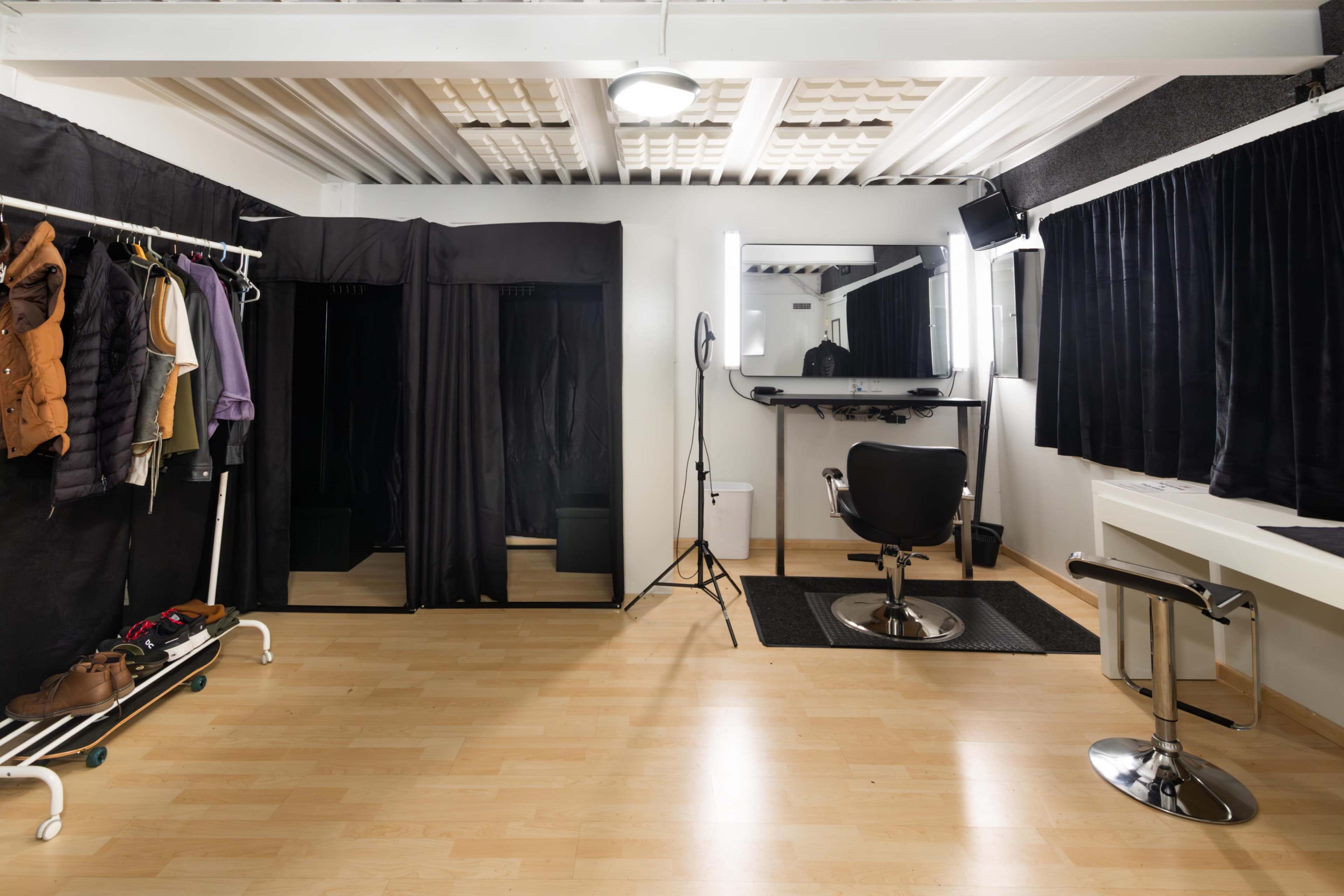 The image features a minimalist hair salon with a black stylist chair, a large mirror, a hanging light, and clothing racks against the walls.