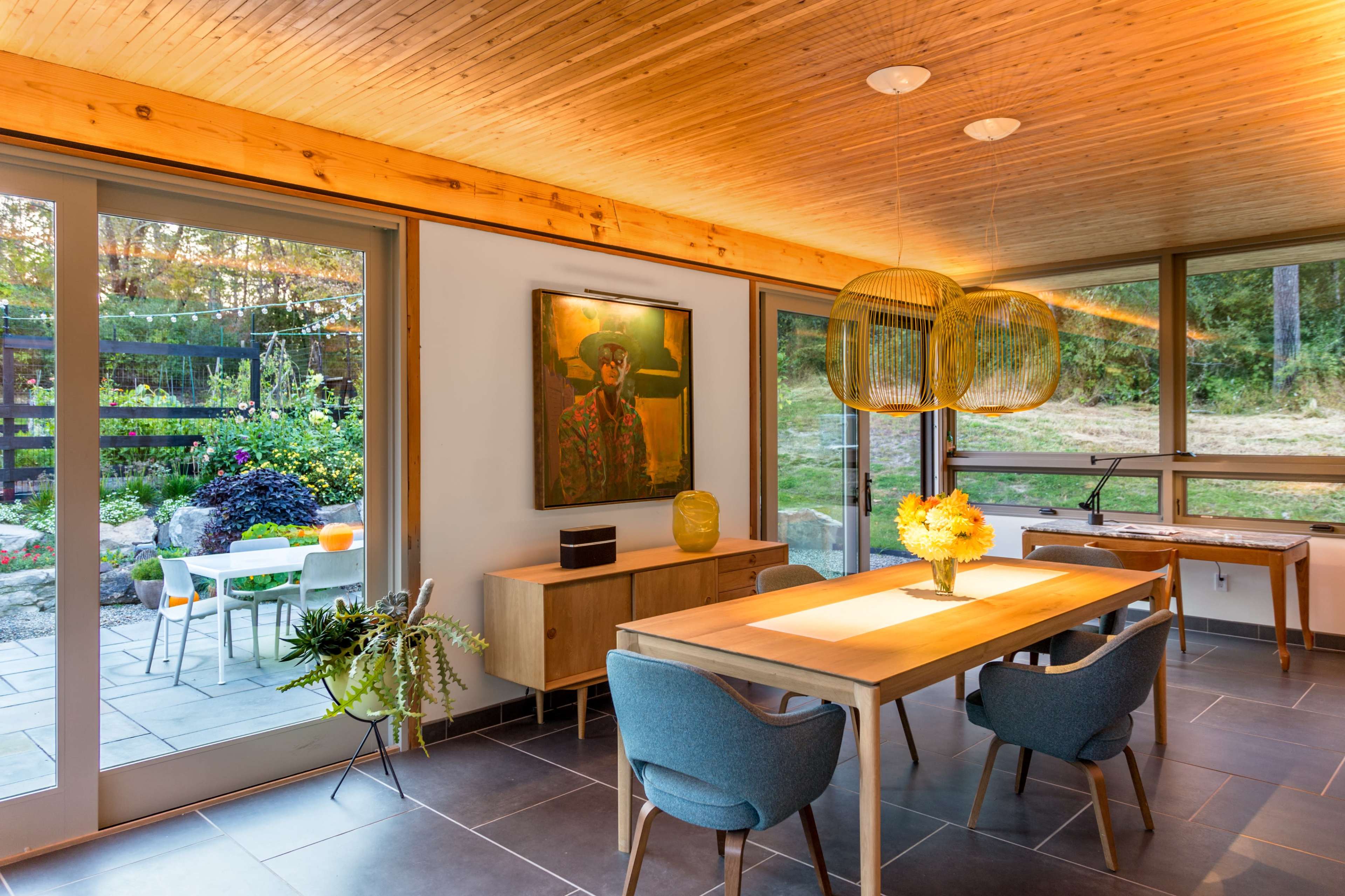 The airy dining area features a wooden table surrounded by blue chairs, with large windows overlooking a landscaped garden.