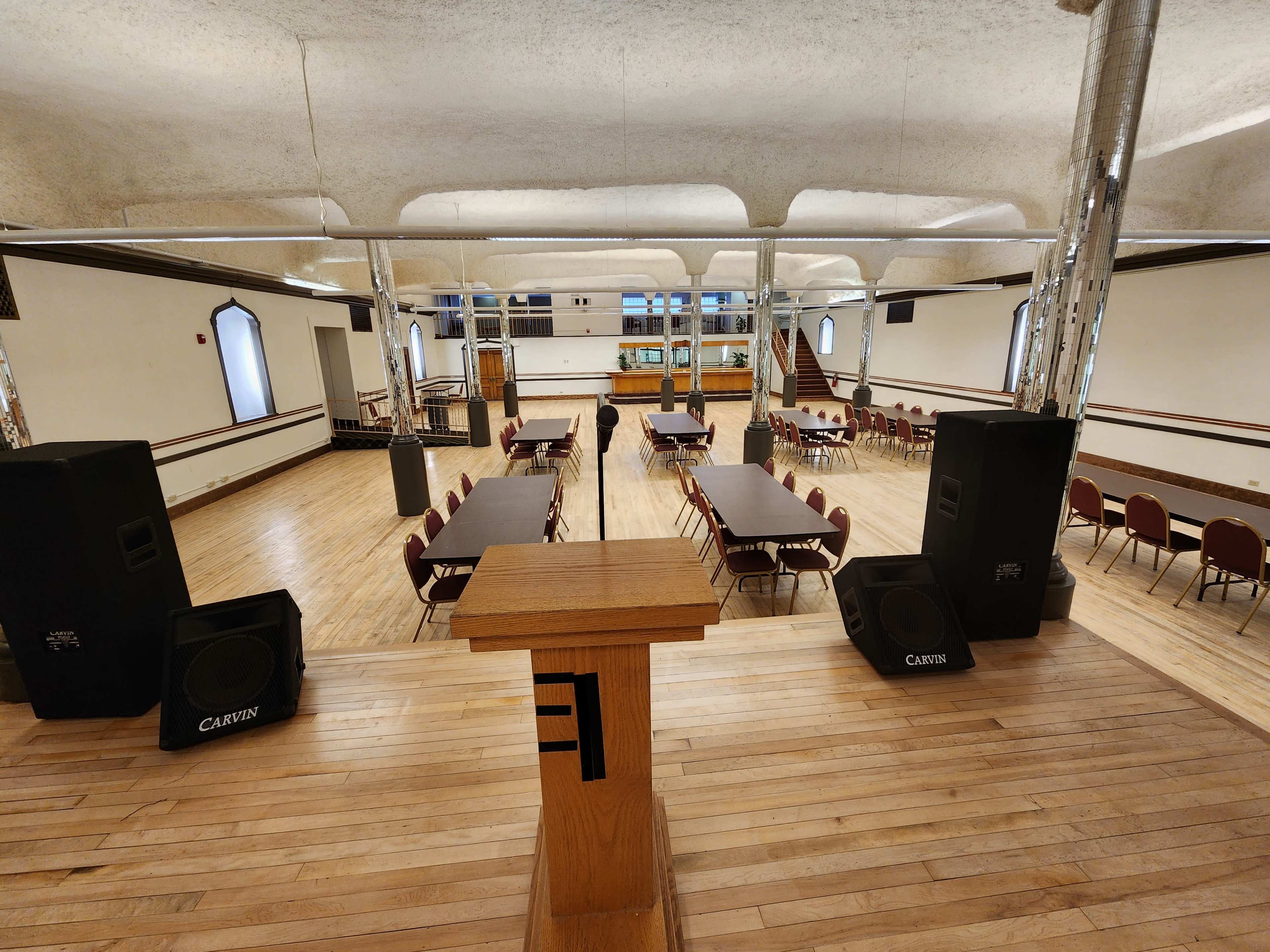 A spacious hall set up with several tables and chairs, featuring a podium at the front and sound equipment on either side.
