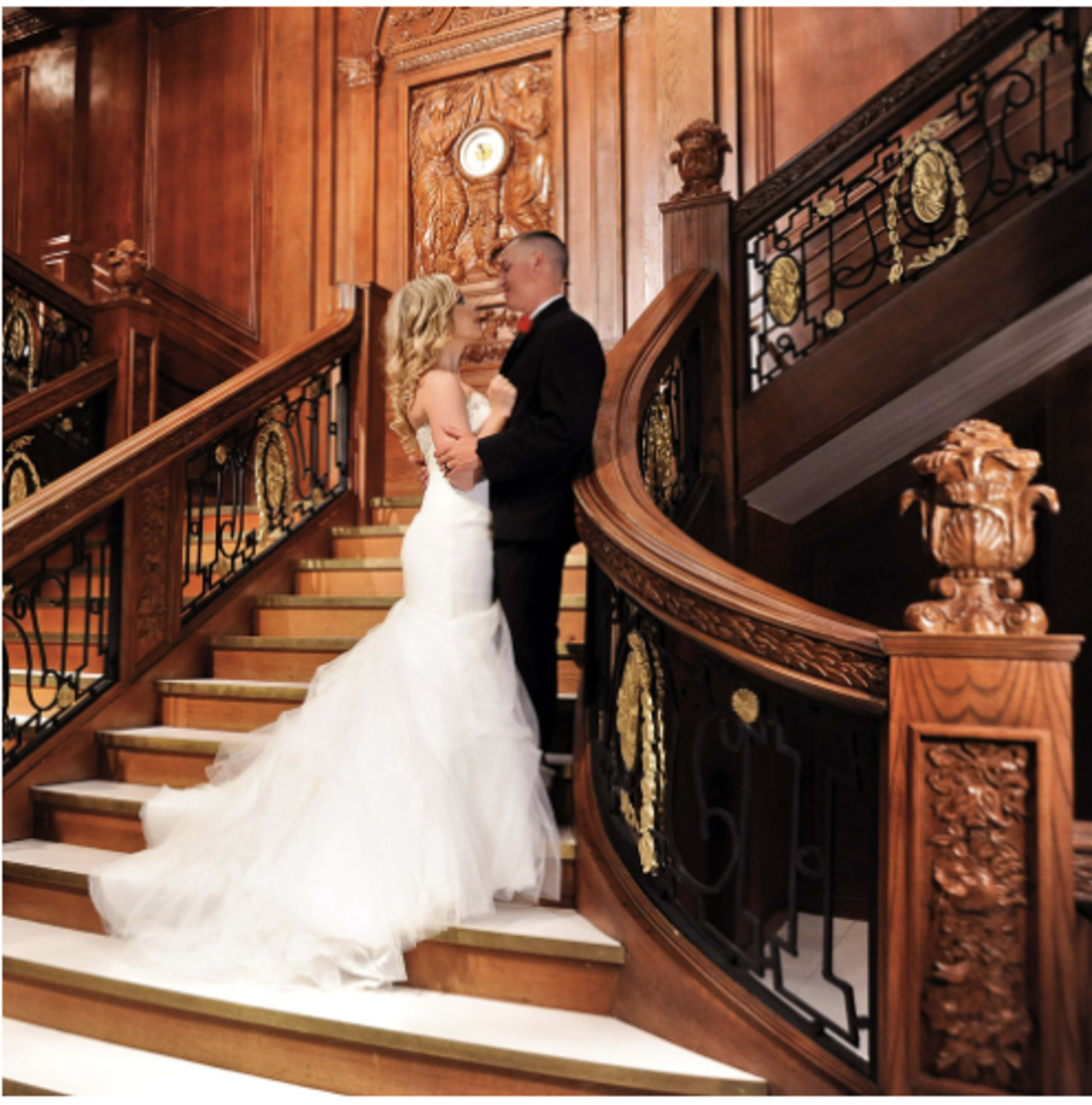 A couple stands on an elegant staircase adorned with wooden railings and intricate carvings.
