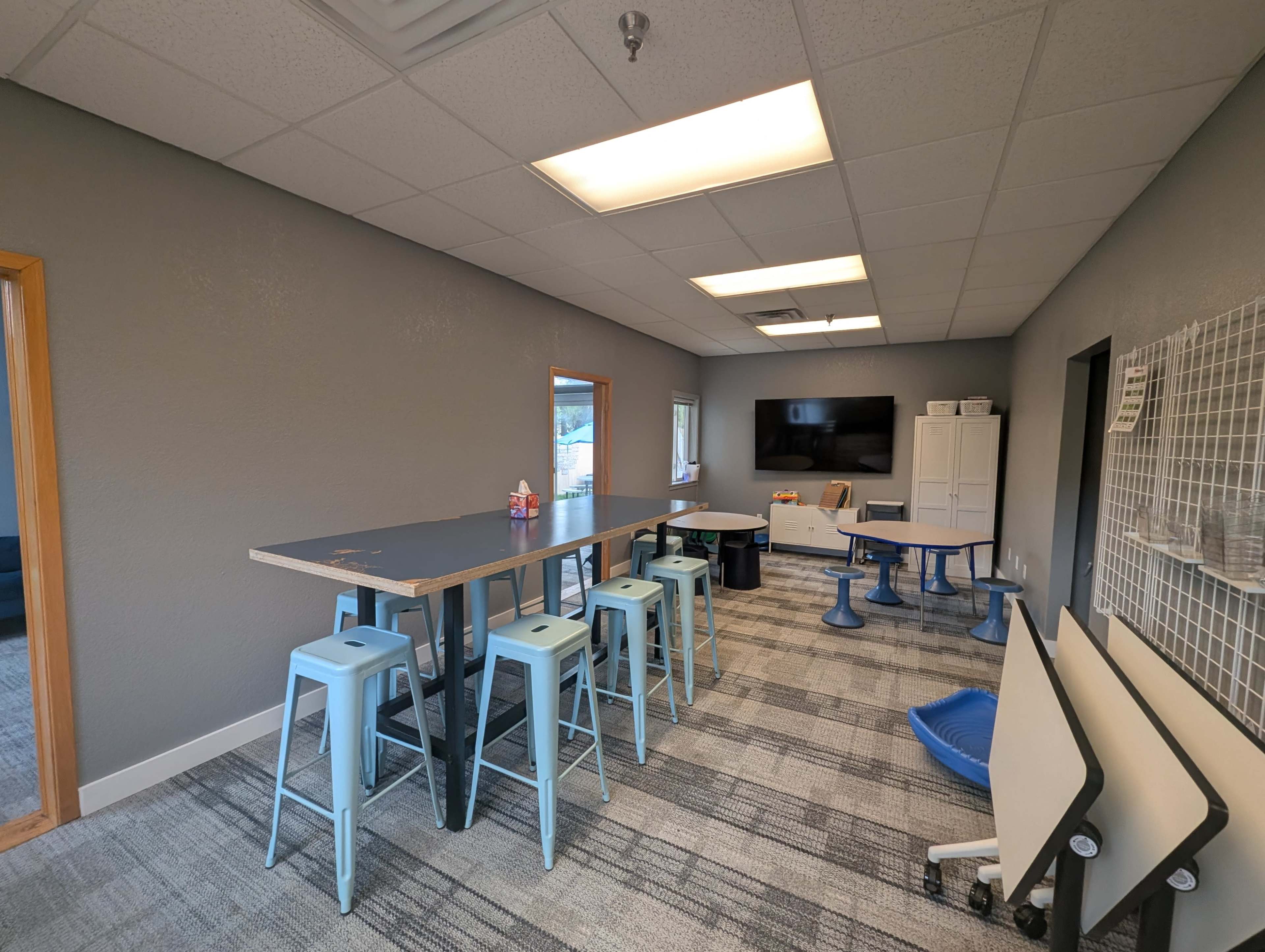 The image shows a modern workspace with a long table, stools, and various office furniture arranged neatly in a well-lit room.