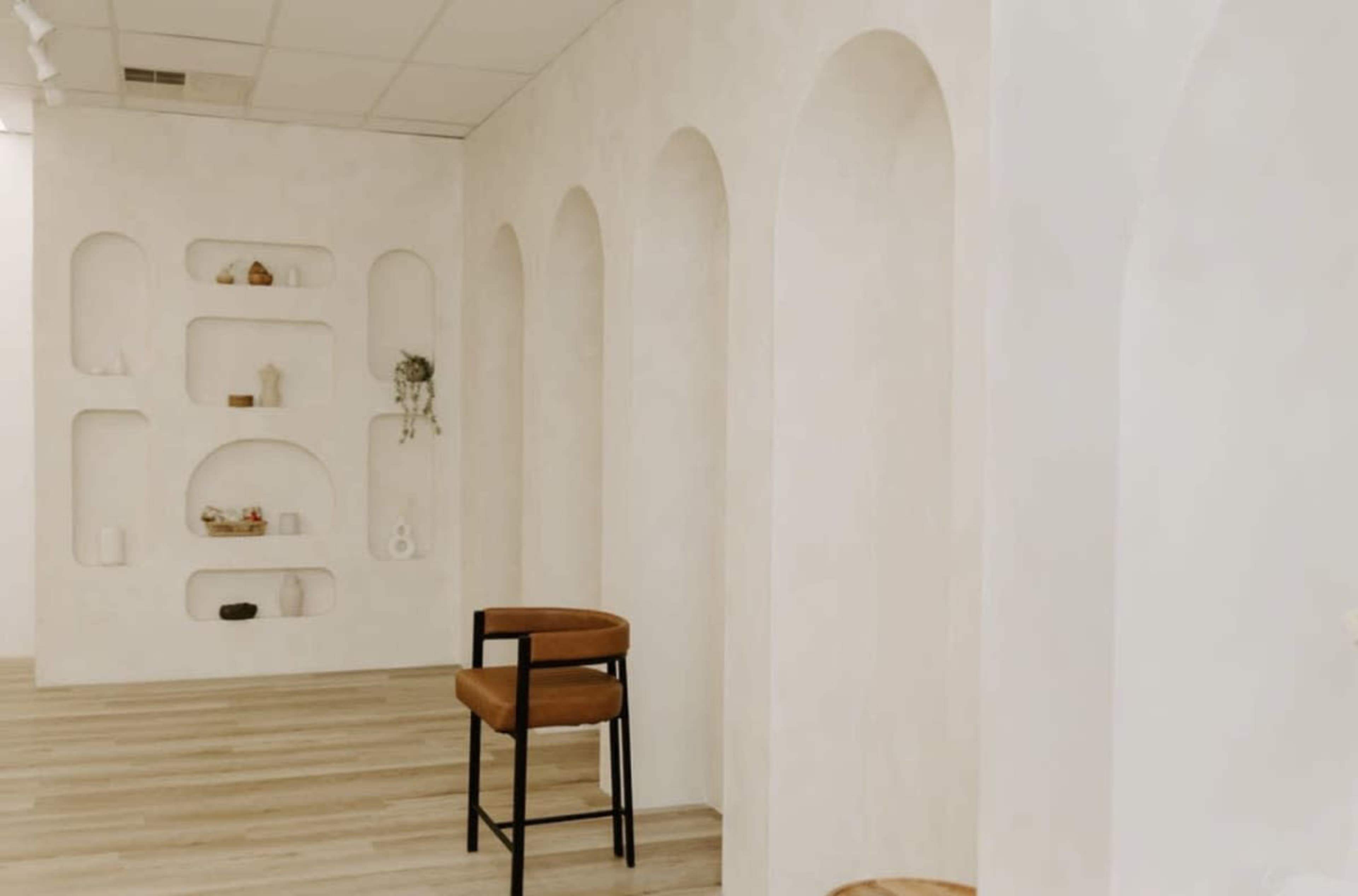 The image shows a minimalist interior space with arched niches on the walls and a wooden stool in the foreground.