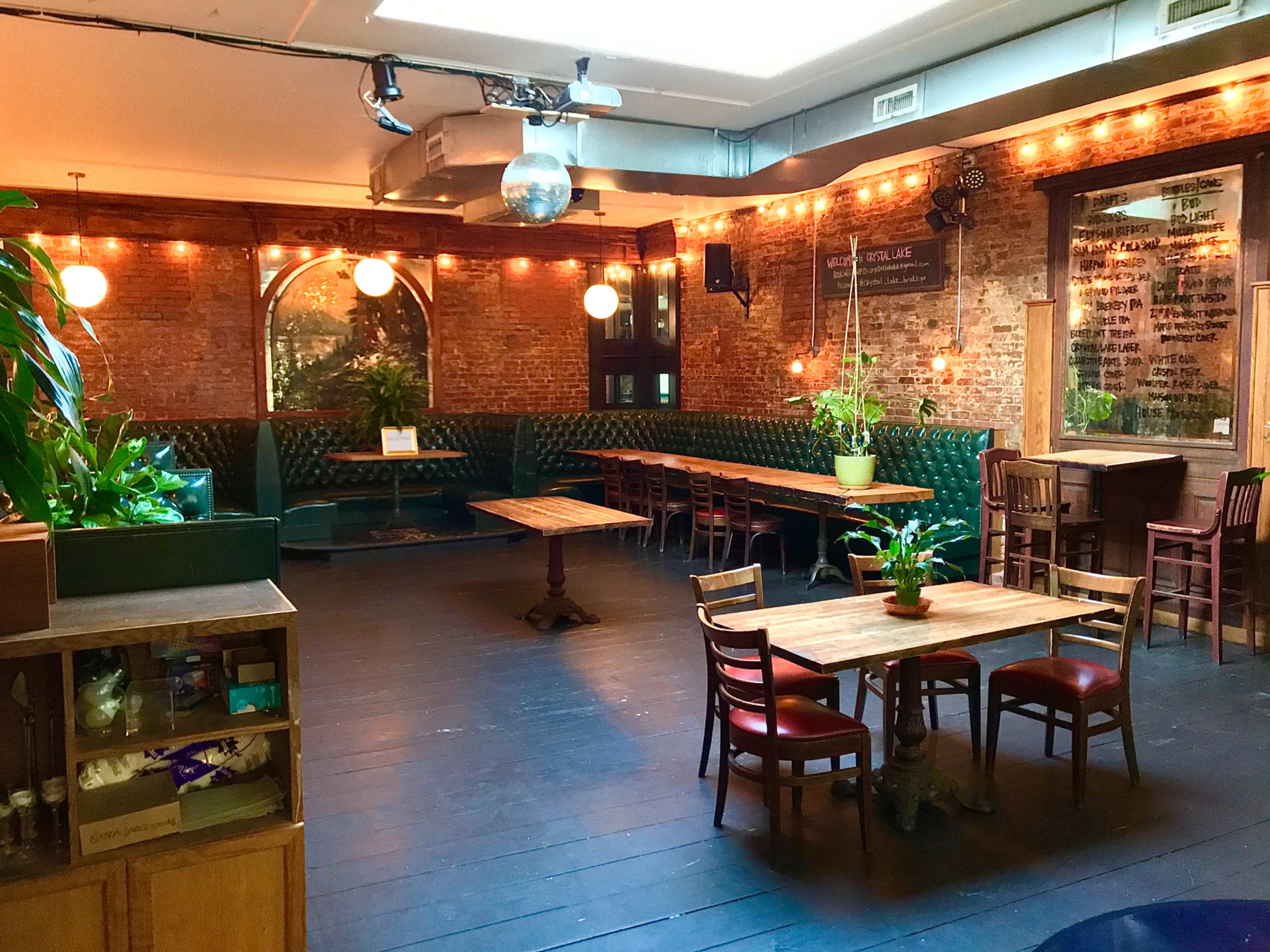 A spacious restaurant interior with dark wooden floors, green upholstered seating, and various tables arranged around the room, illuminated by warm lighting and plants.