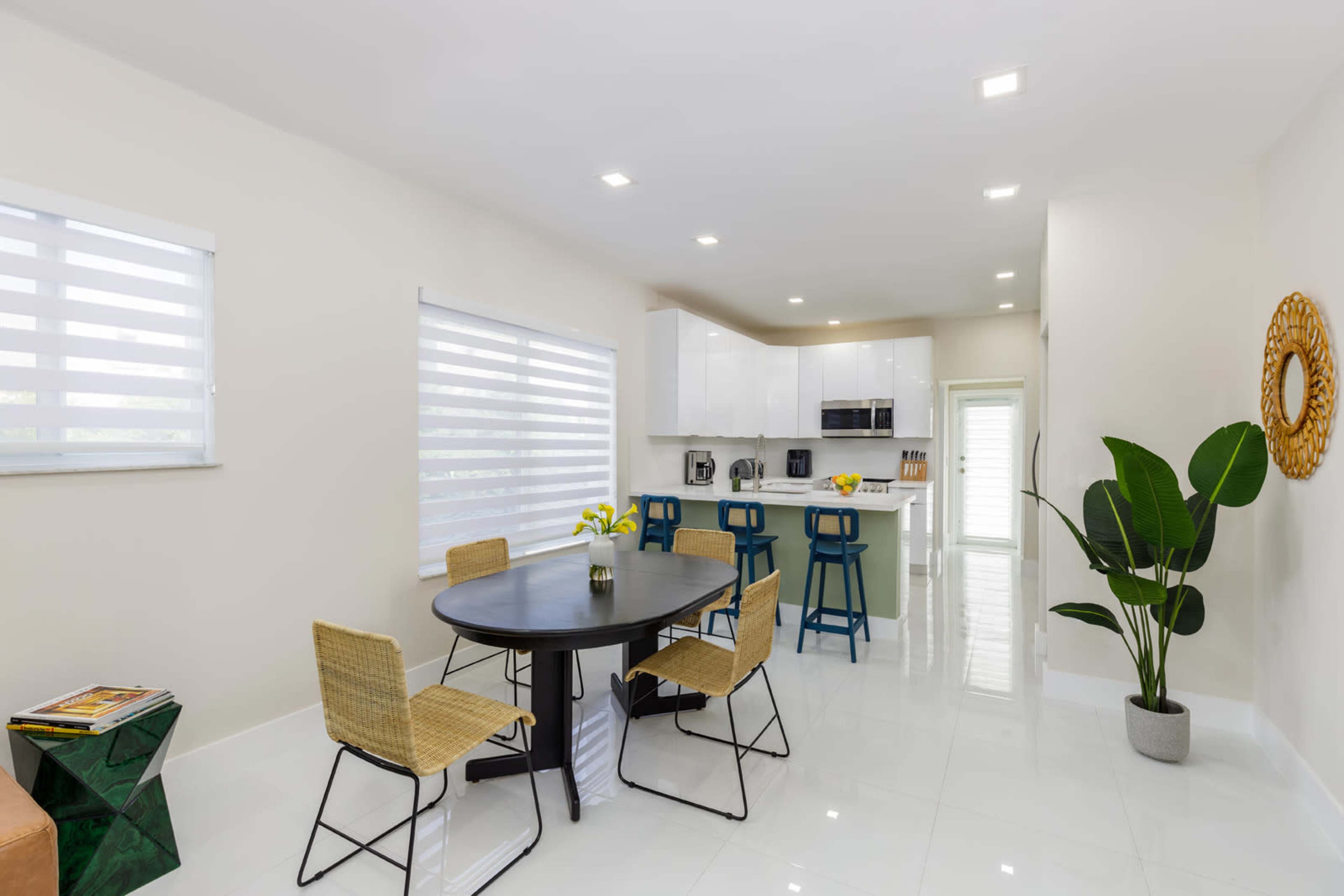 A modern kitchen and dining area features a round black table with four chairs, a sleek kitchen with white cabinets, and a large window with blinds.