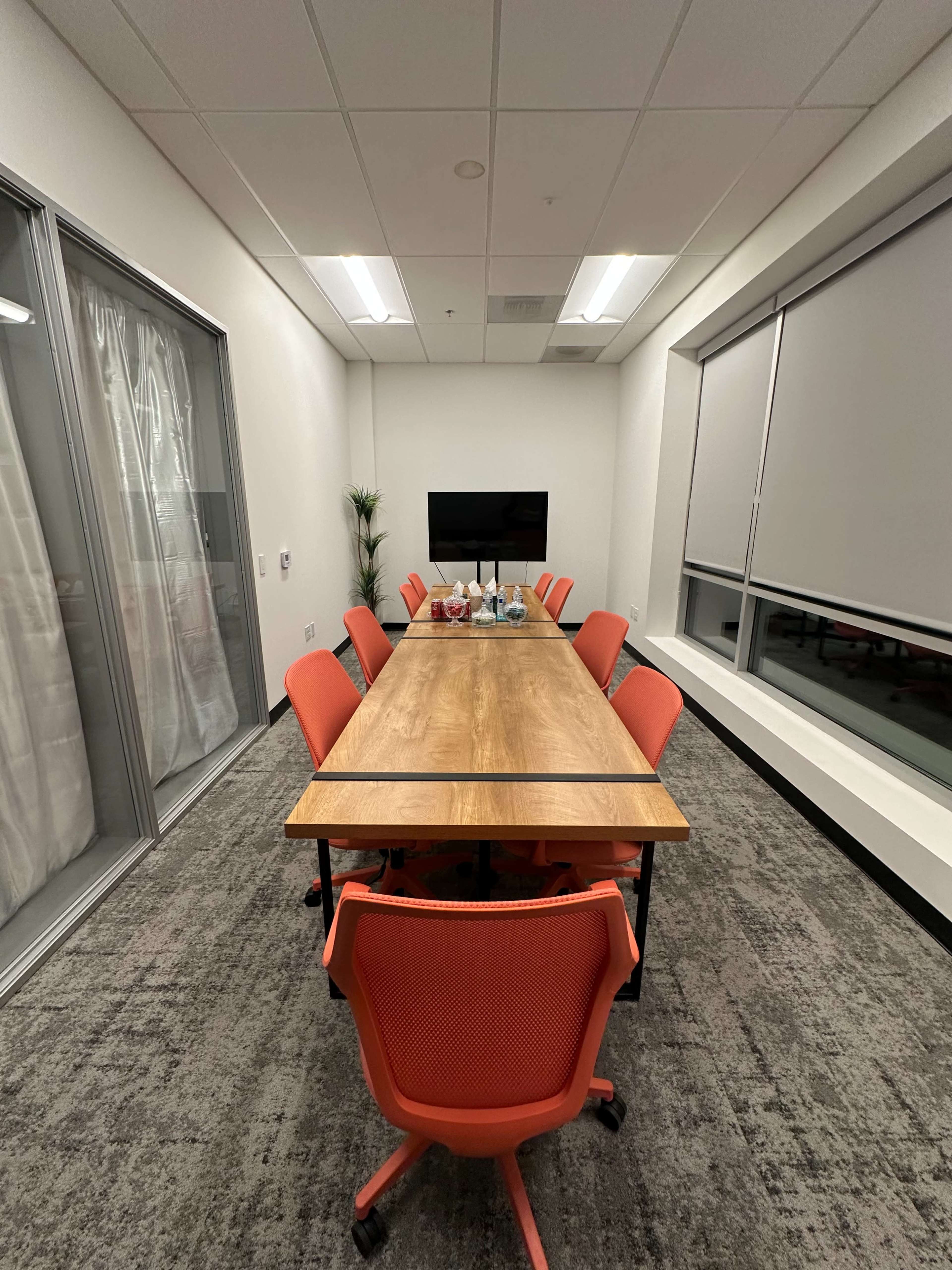 A conference room features a long wooden table surrounded by eight orange chairs, with large windows on one side and a television mounted on the wall.