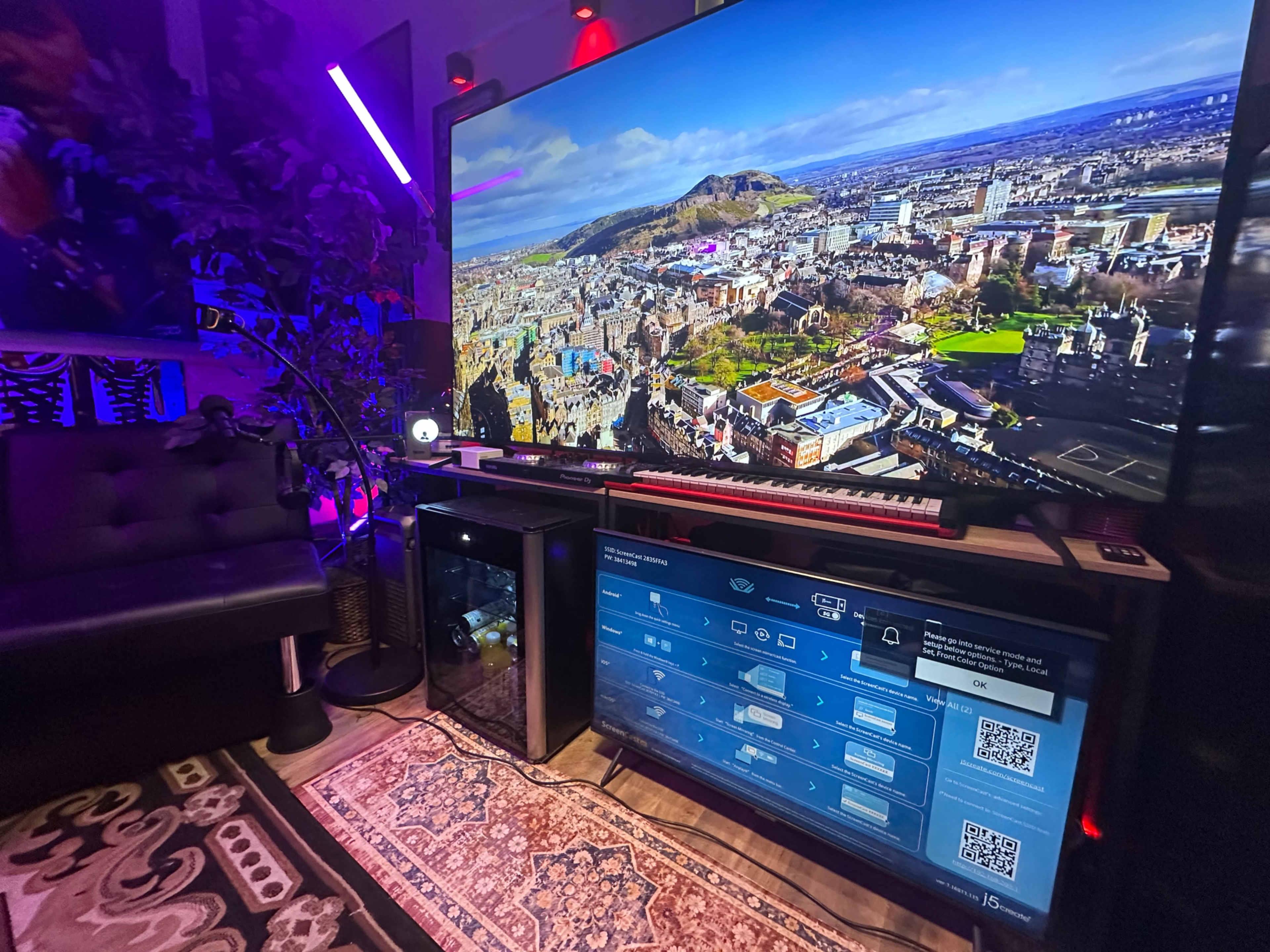 A large TV displaying an aerial view of a city is positioned near a black couch and a monitor with visible settings and QR codes, all set in a dimly lit room with decorative lighting.