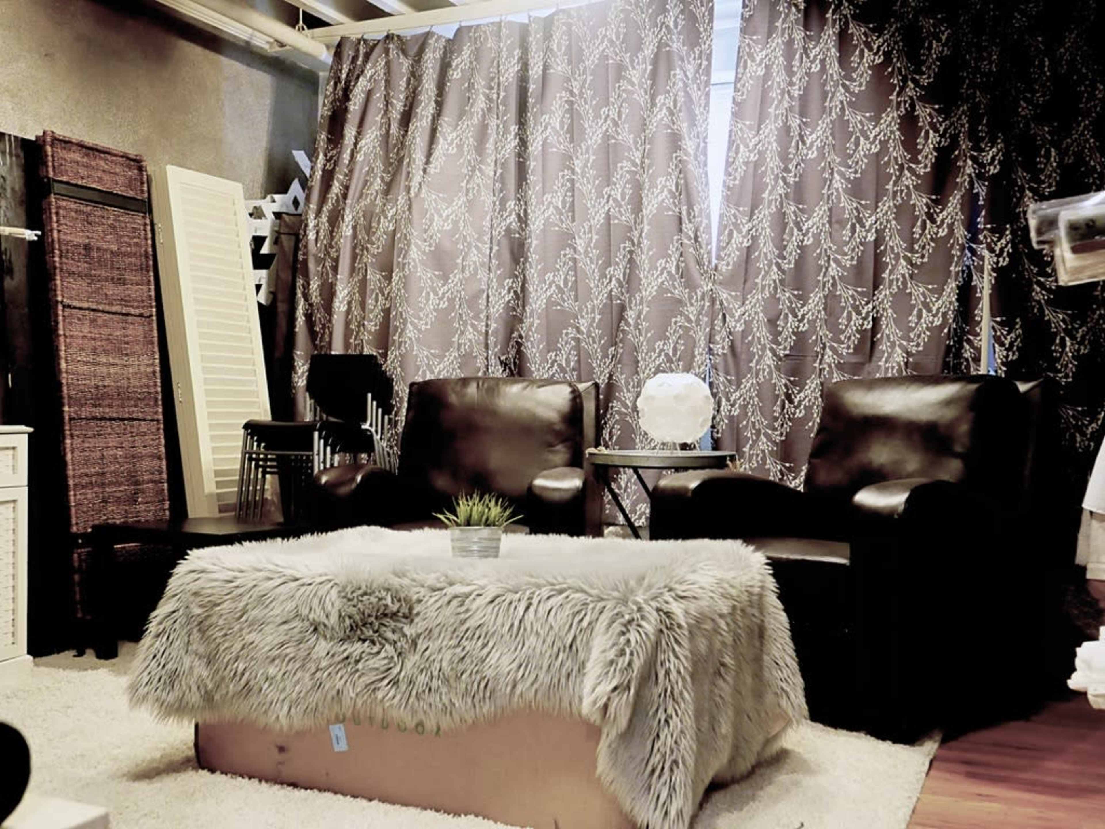 The image shows a cozy living room scene with two black chairs, a decorative table, and a fluffy throw covering a coffee table, surrounded by patterned curtains.