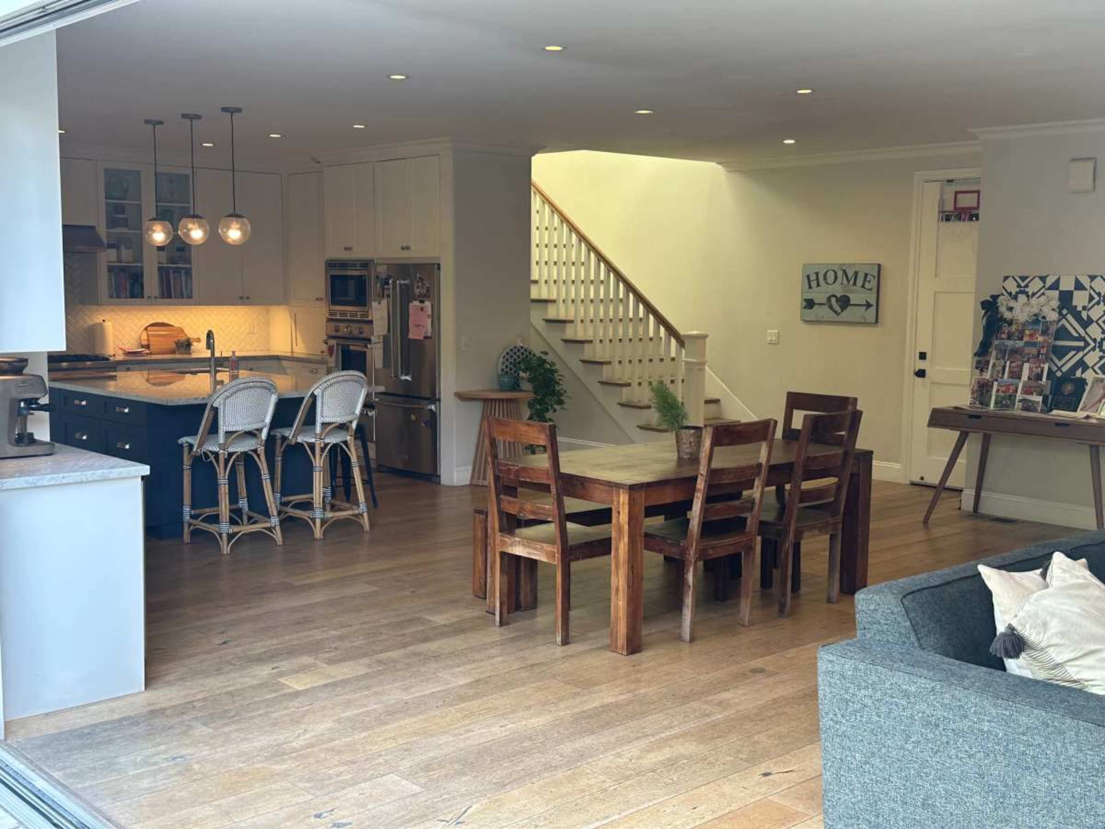 The image shows a spacious kitchen and dining area with a wooden table, chairs, and an open staircase in a well-lit room.