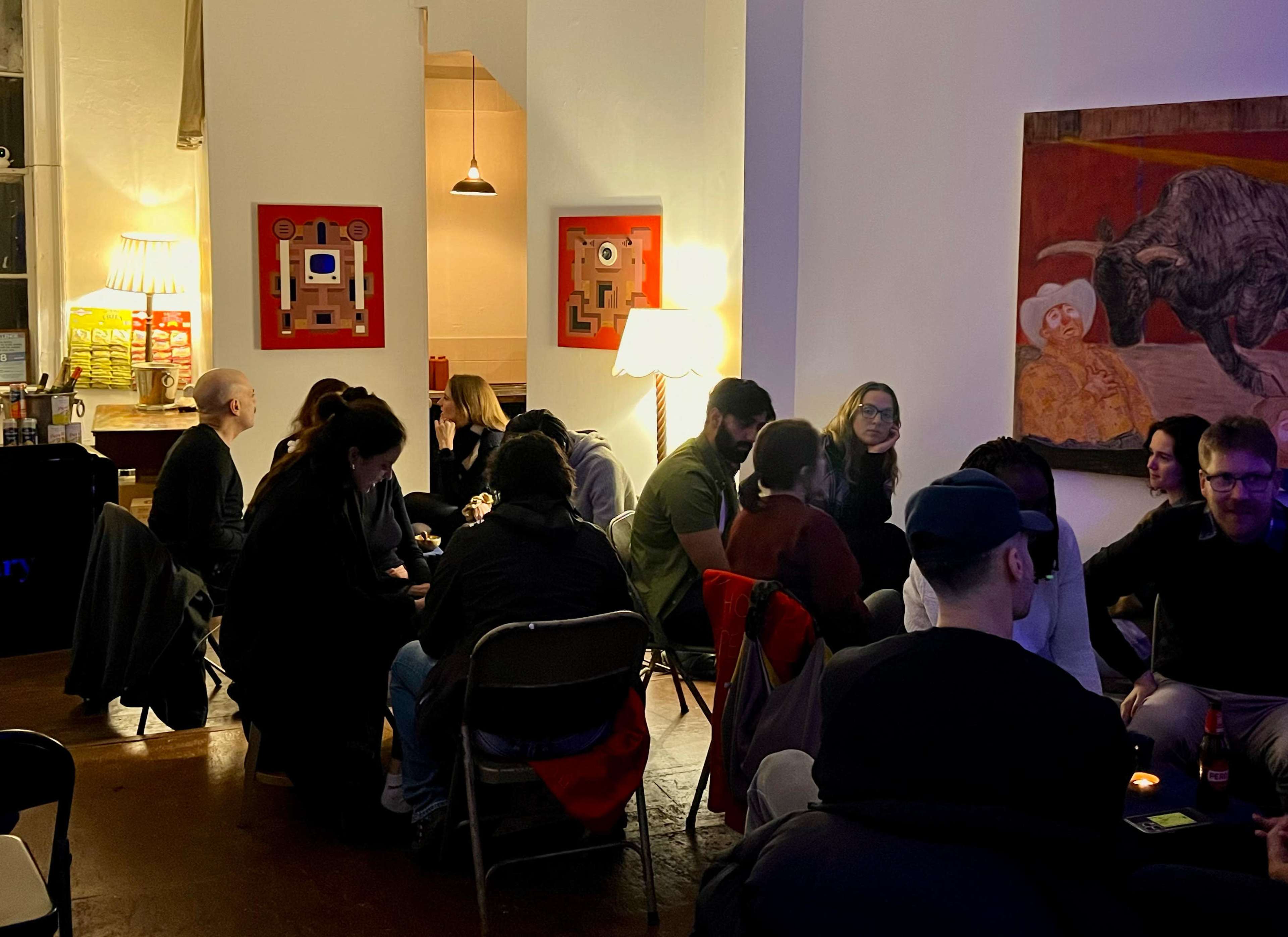 A group of people are seated in a warmly lit room, engaged in conversation, with artwork on the walls and chairs arranged in a circle.