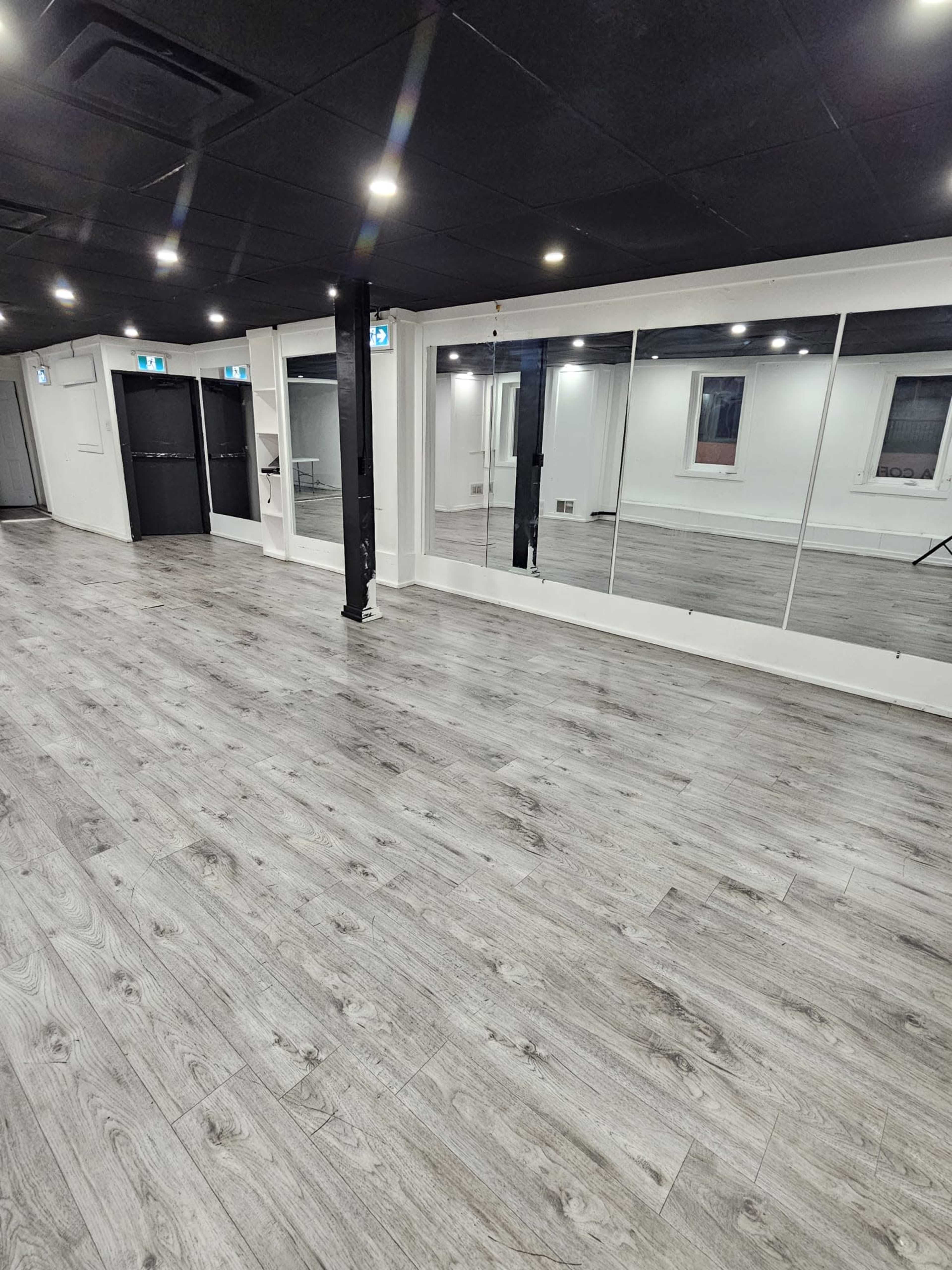 The image shows an empty interior room with gray wood flooring, large mirrors on one wall, and bright overhead lighting.