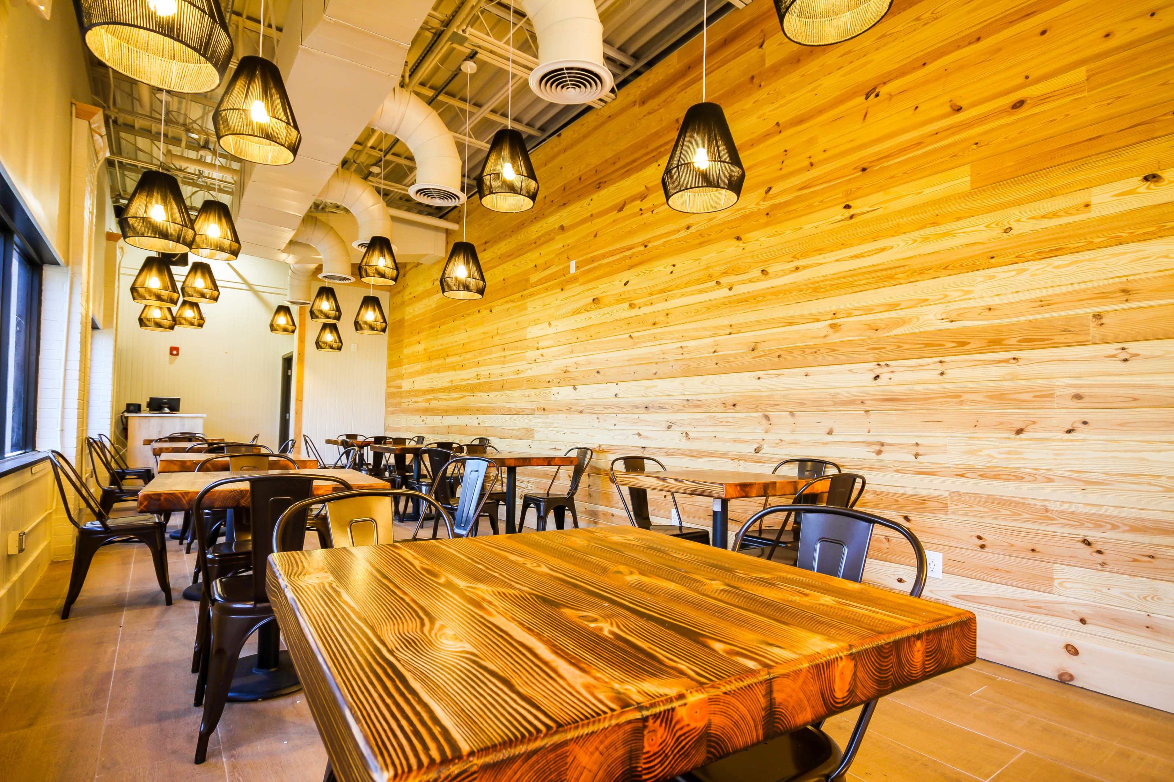 A bright dining area features wooden tables and black chairs against a backdrop of a light wood-paneled wall with pendant lighting.