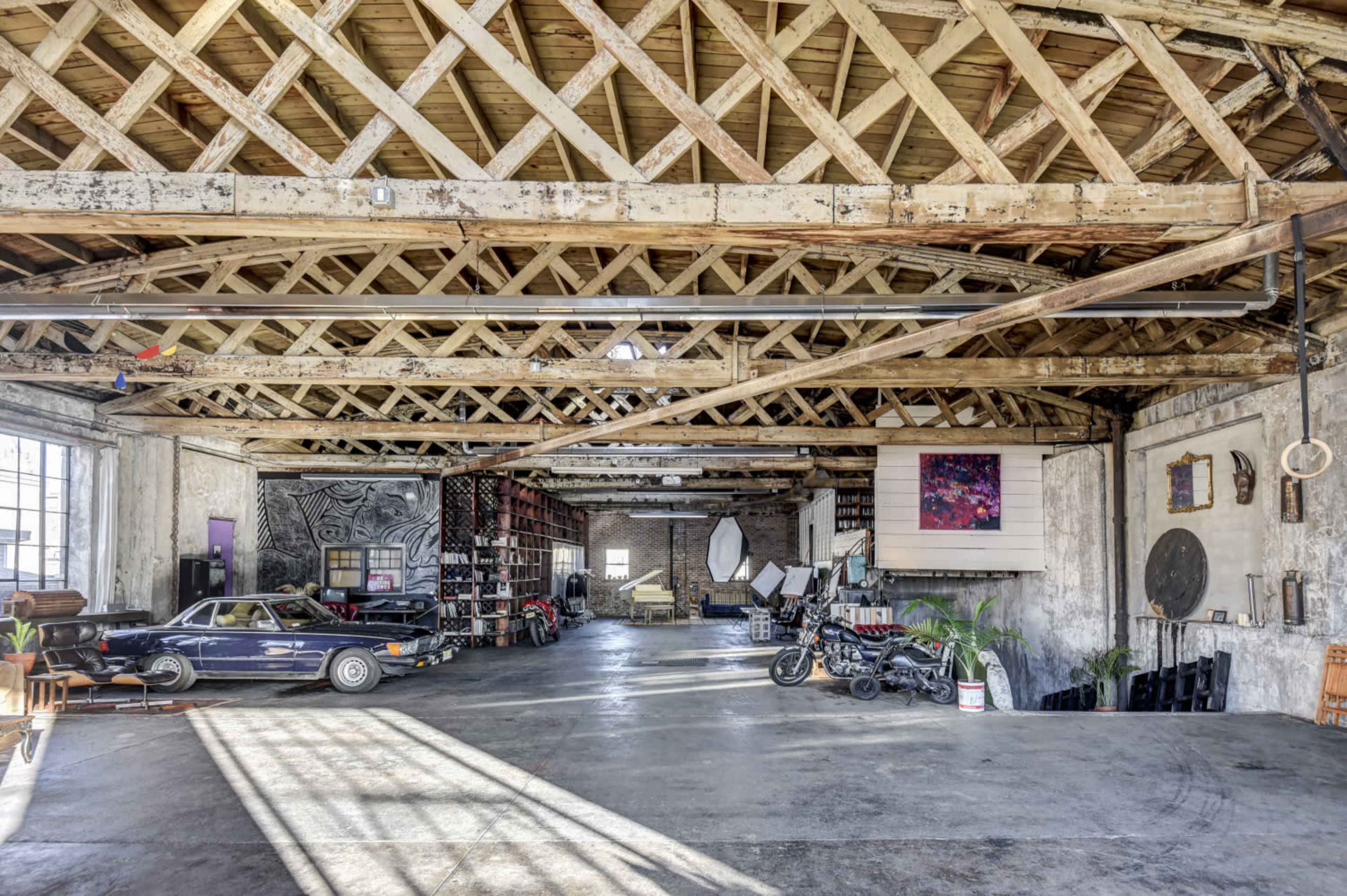 A spacious, industrial-style warehouse interior with wooden beams, various furniture items, a parked car, and artistic decorations on the walls.