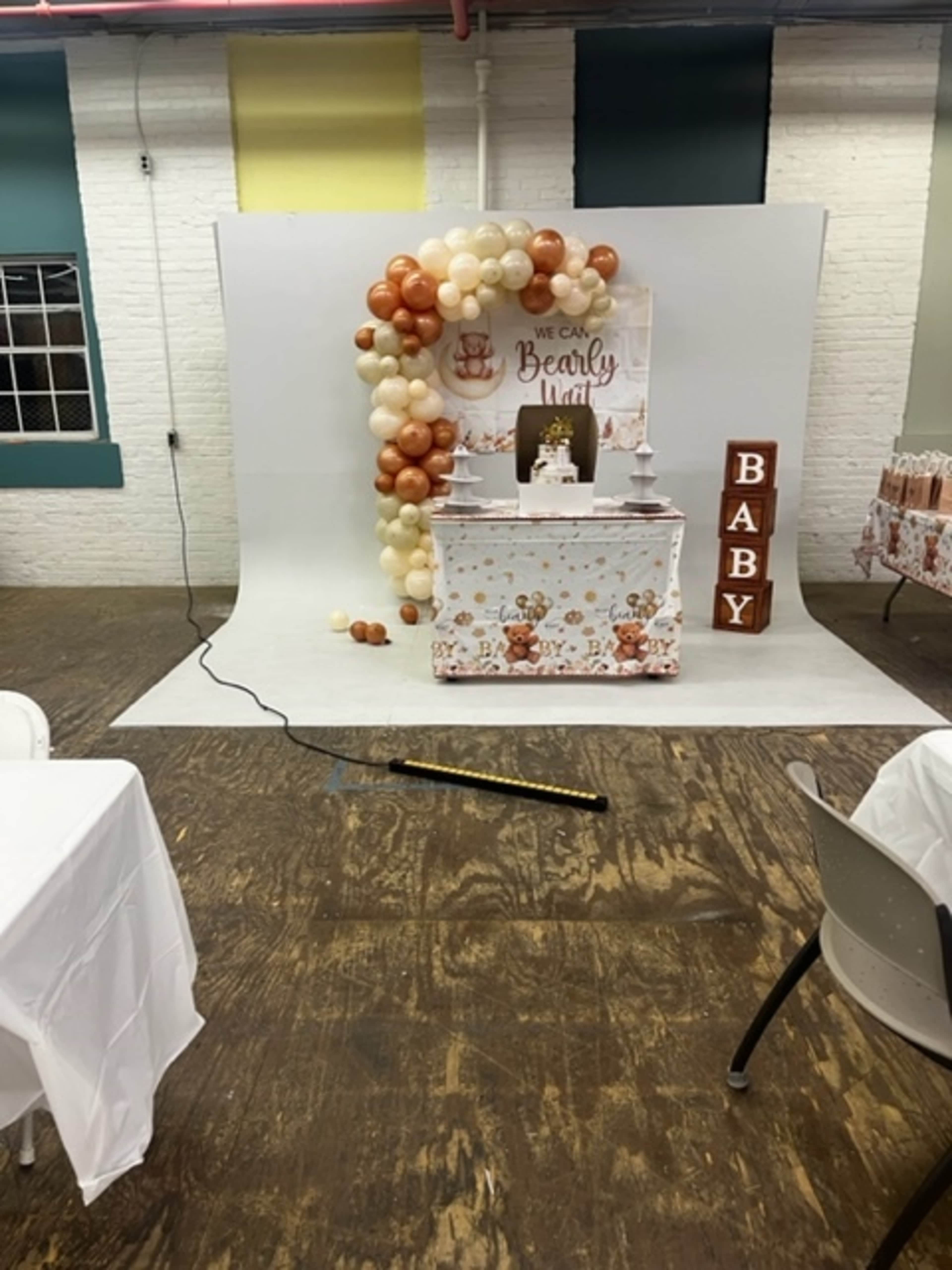 The image shows a decorated event space with a backdrop of balloons, a small table with a cake, and "BABY" letter blocks arranged for a baby shower celebration.