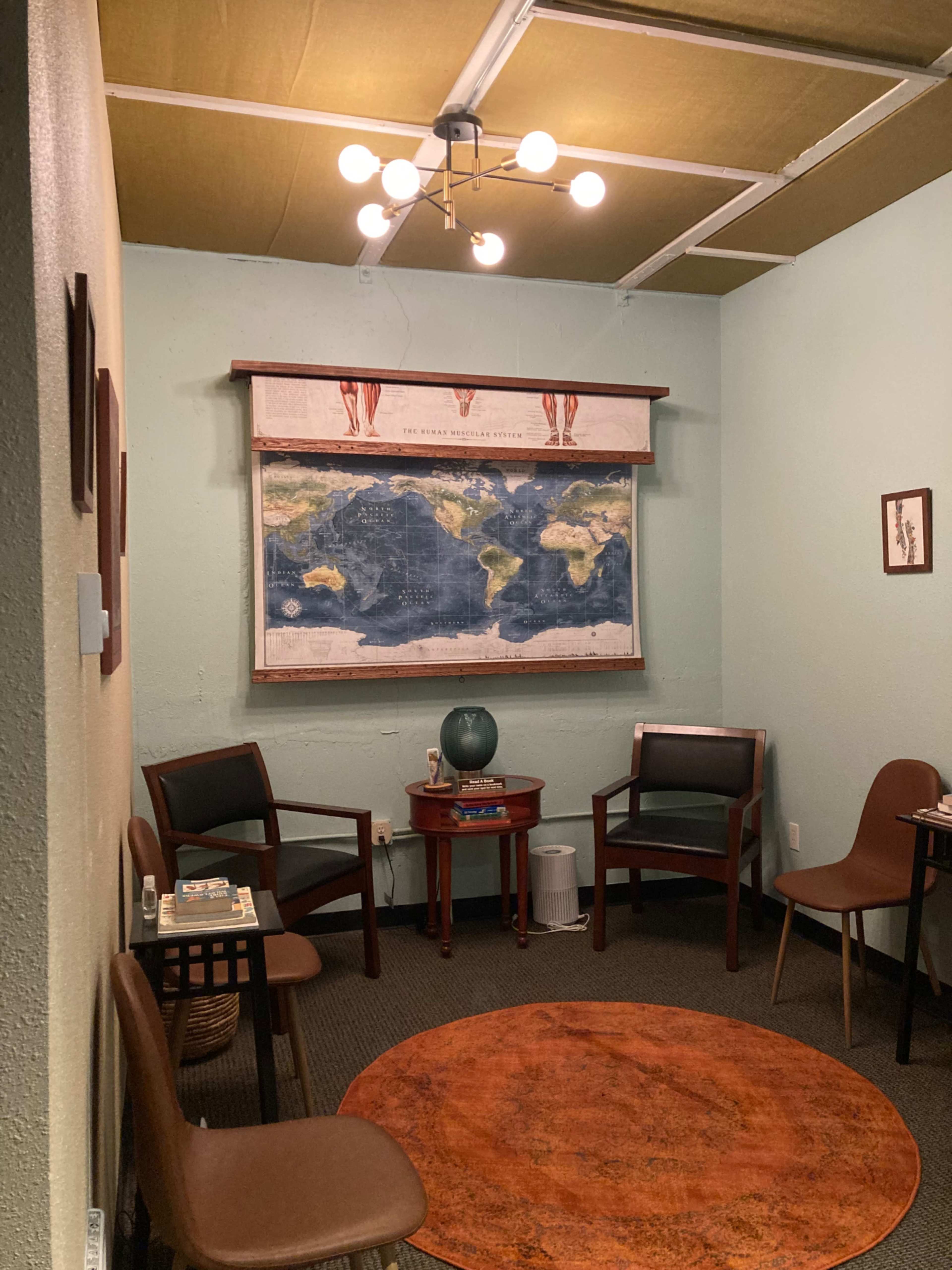 A small room with a round orange rug, four chairs, a wooden table, and a world map mounted on the wall.