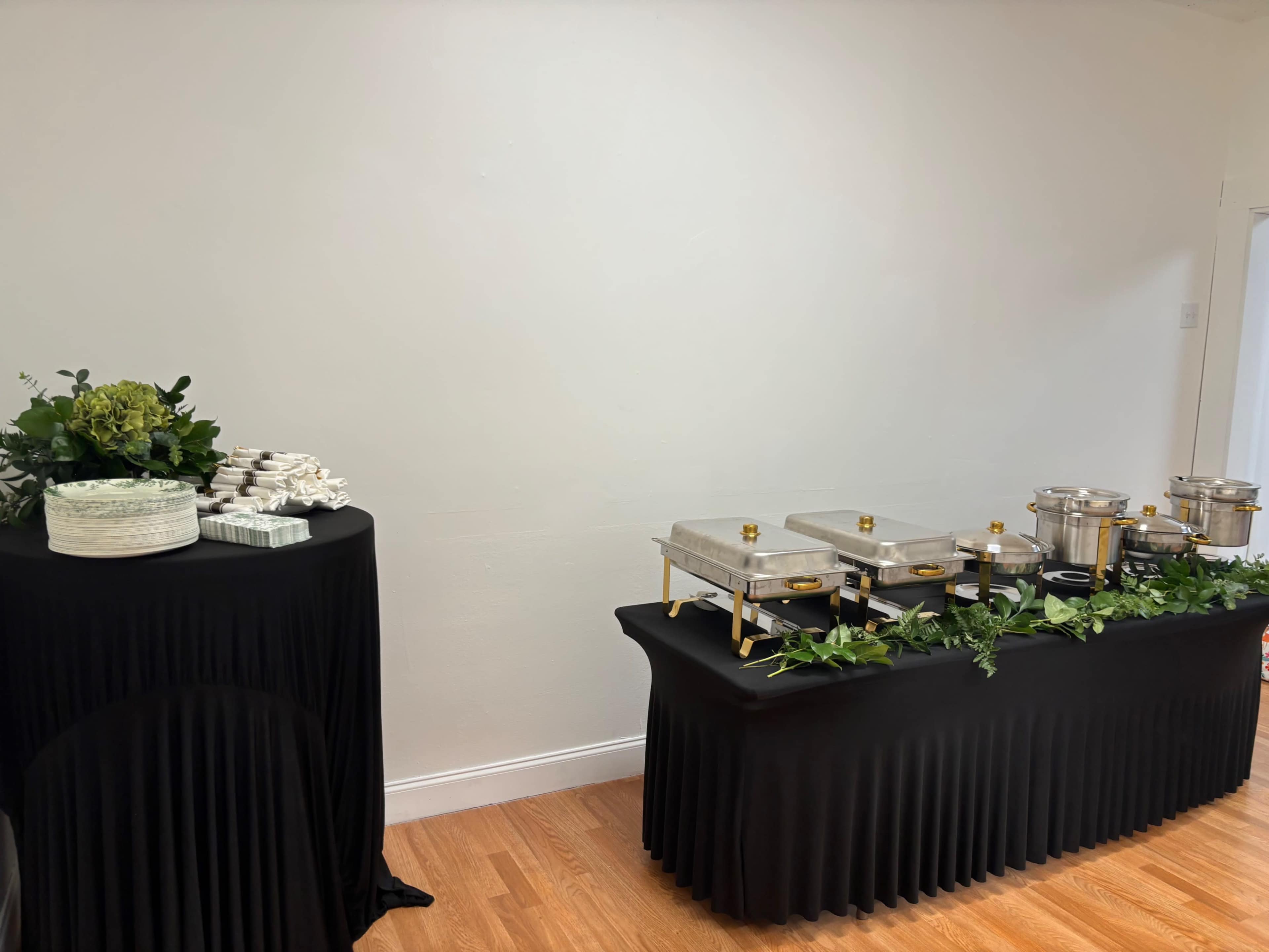 A black tablecloth covers a buffet display featuring chafing dishes and a selection of plates and napkins arranged on a separate table.