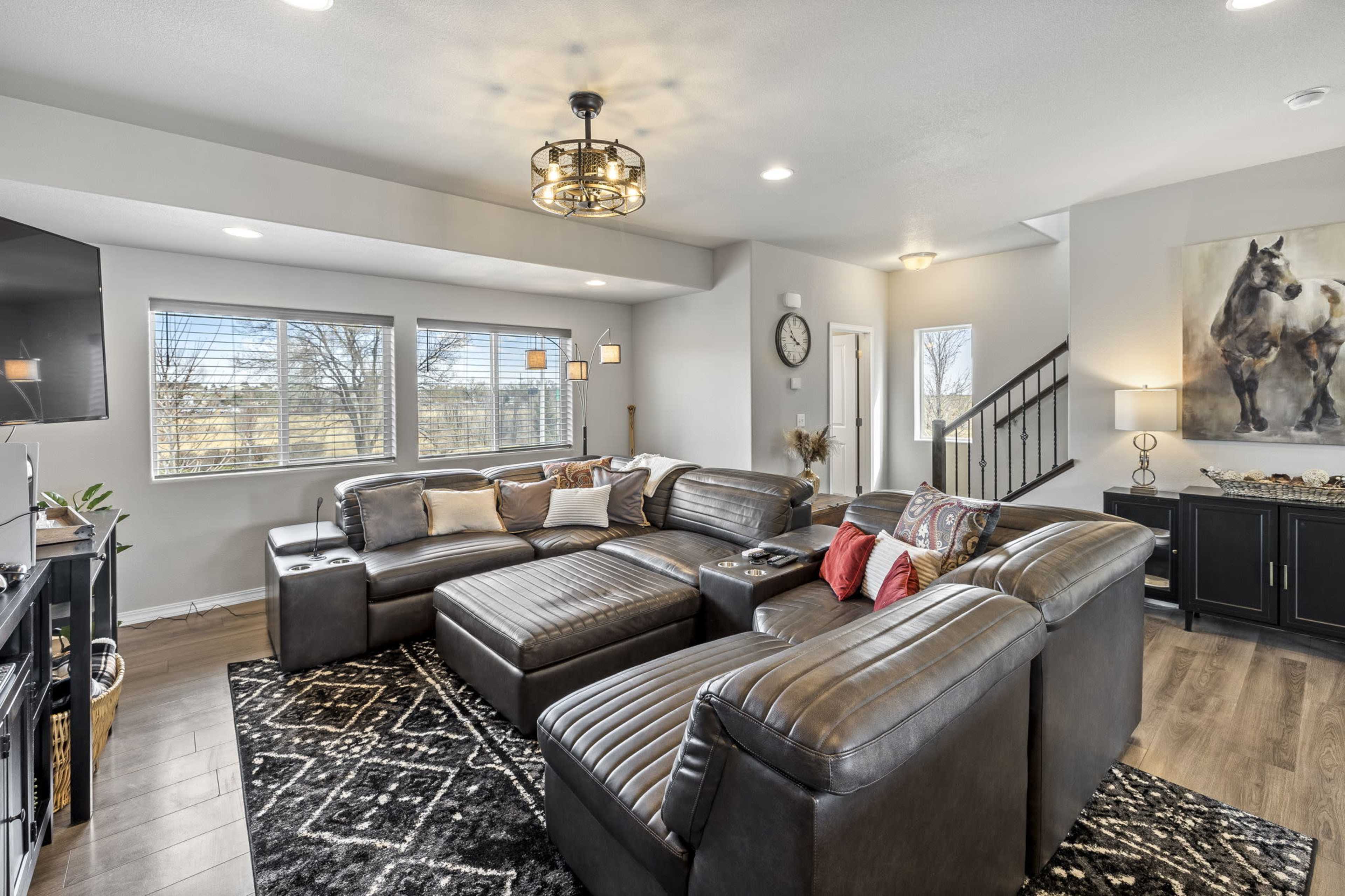 A spacious living room with a large, dark sectional sofa, a coffee table, and windows allowing natural light, featuring a modern chandelier and a painting of a horse on the wall.